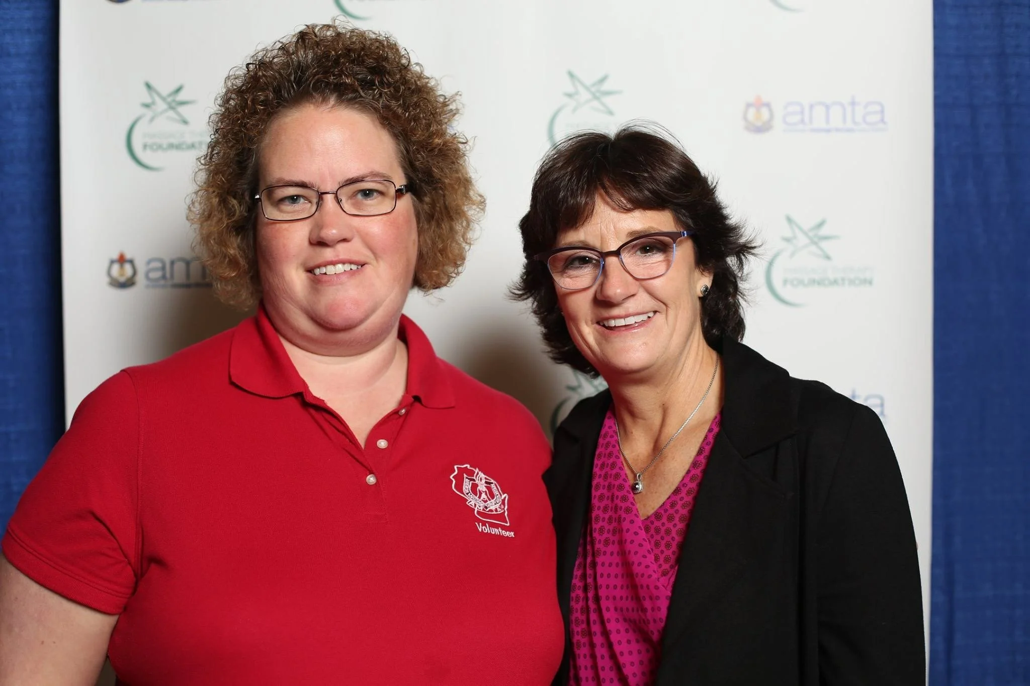 Two women smiling, standing close together in front of a backdrop with logos, one wearing a red polo shirt with a volunteer emblem and the other in a black jacket and purple polka-dot blouse.
