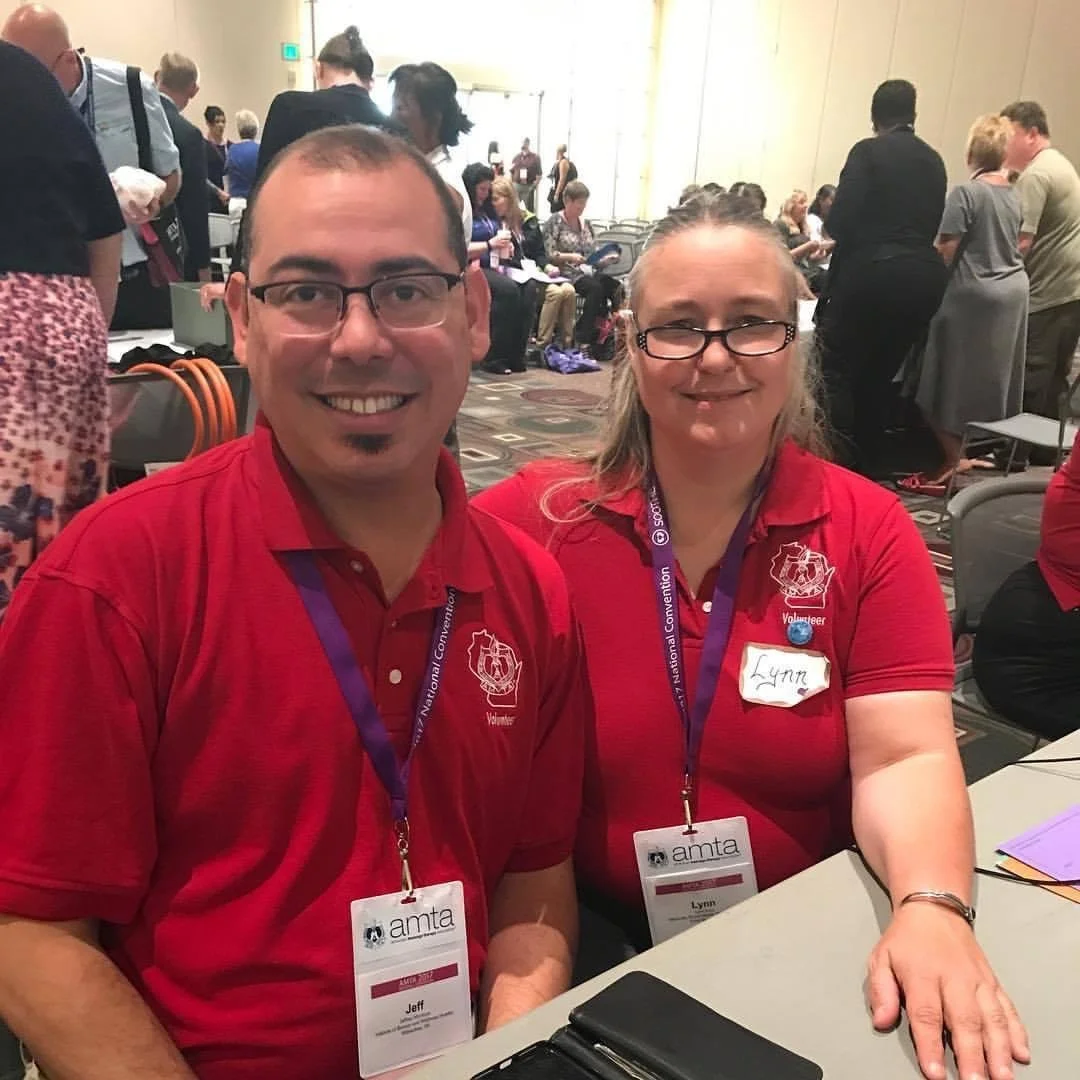 Two volunteers wearing red shirts with AMTA badges sitting at a table at a conference, with many people in the background.