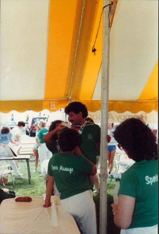 People gathered under a yellow and white striped tent at an outdoor event, with some wearing green shirts that read 'Santa Manage' and 'Sport'.