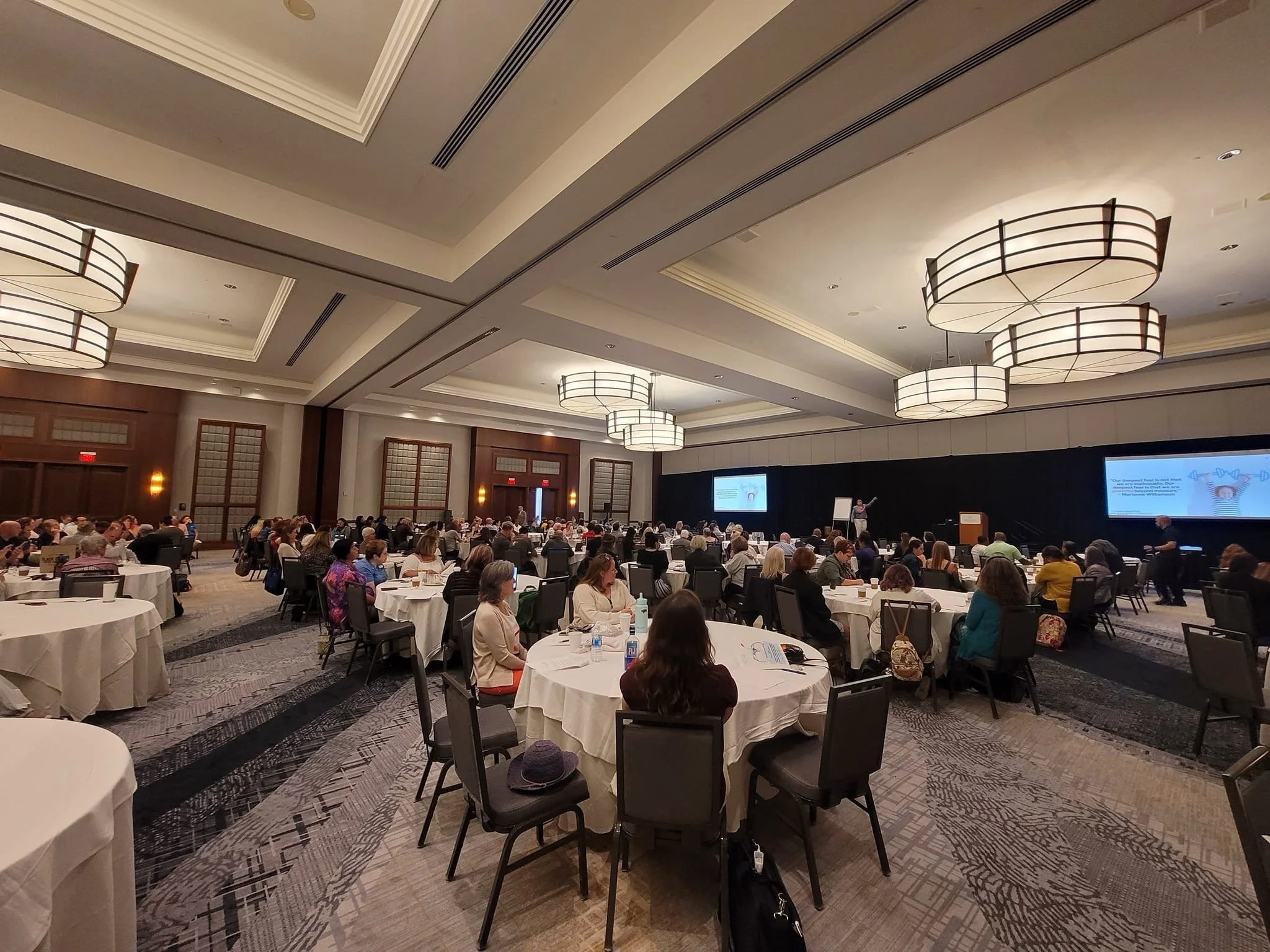 A large conference room filled with round tables and attendees listening to a speaker at the front of the room. Large hanging lights and high ceilings.