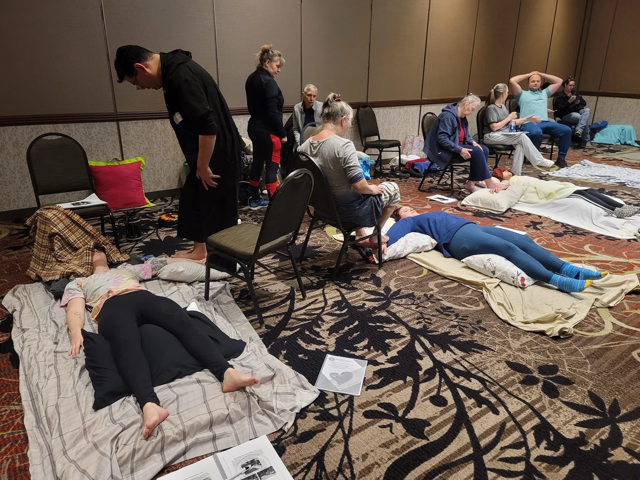 People participating in a CPR training session in a conference room. Several are lying on mats practicing CPR, while others observe or wait their turn. Instructional materials and personal belongings are scattered around the room.