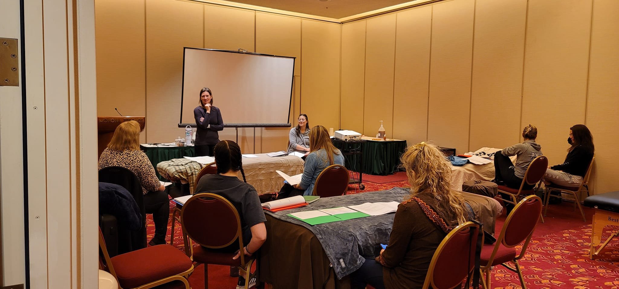 A group of women attending a workshop or seminar in a conference room, with one woman standing and speaking at the front while others sit and listen, some taking notes.