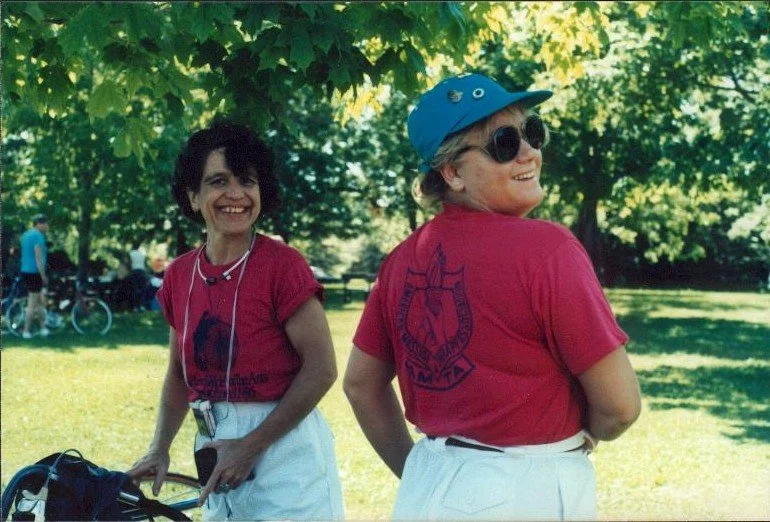 Two women in red shirts and white pants outdoors under a leafy tree, smiling and enjoying a sunny day, with bicycles in the background.