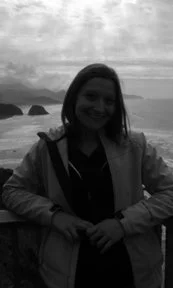 A woman standing outdoors near a body of water with rocks in the background, smiling and wearing a jacket.
