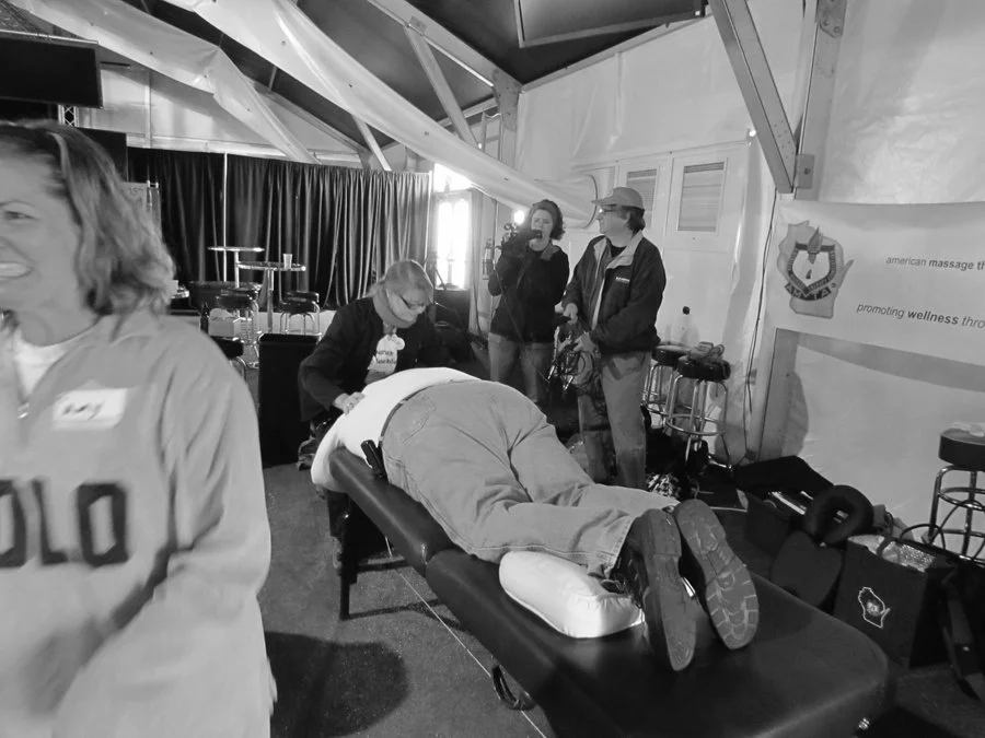 A person lying face down on a massage table while a woman and a man stand nearby. The woman is sitting and smiling, and the man is holding a camera or recording device. There are other people and equipment in the background, suggesting a wellness or 