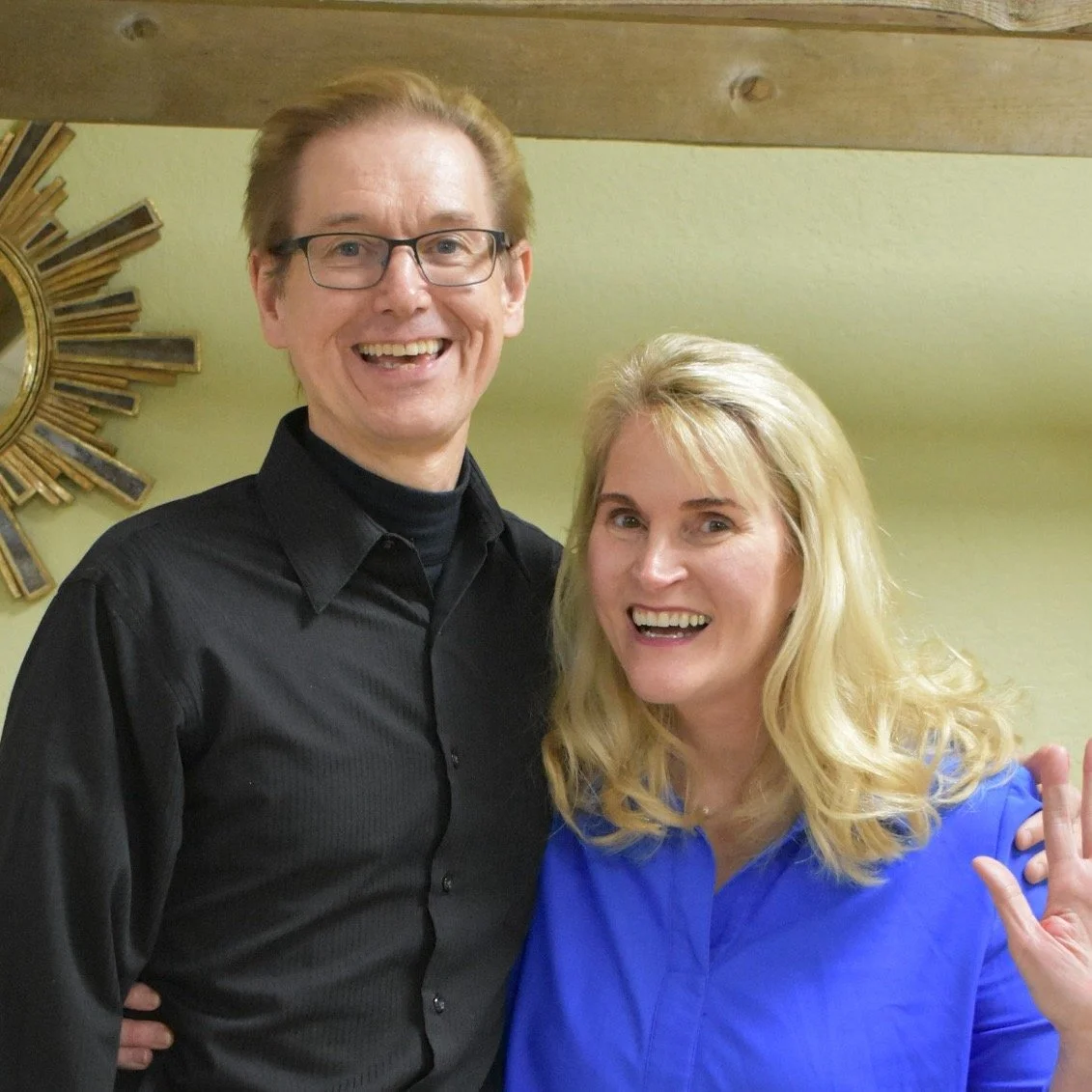A man with glasses and a woman with blonde hair smiling and posing together indoors.