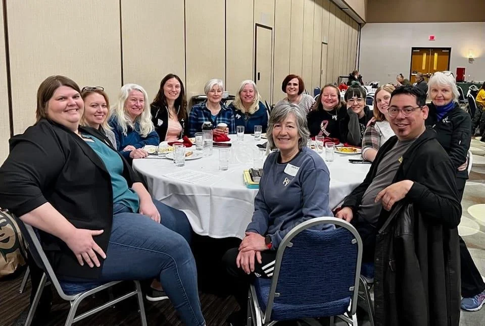 A group of fifteen people gathered around a round table at an indoor event, smiling at the camera, with plates of food and drinks on the table.