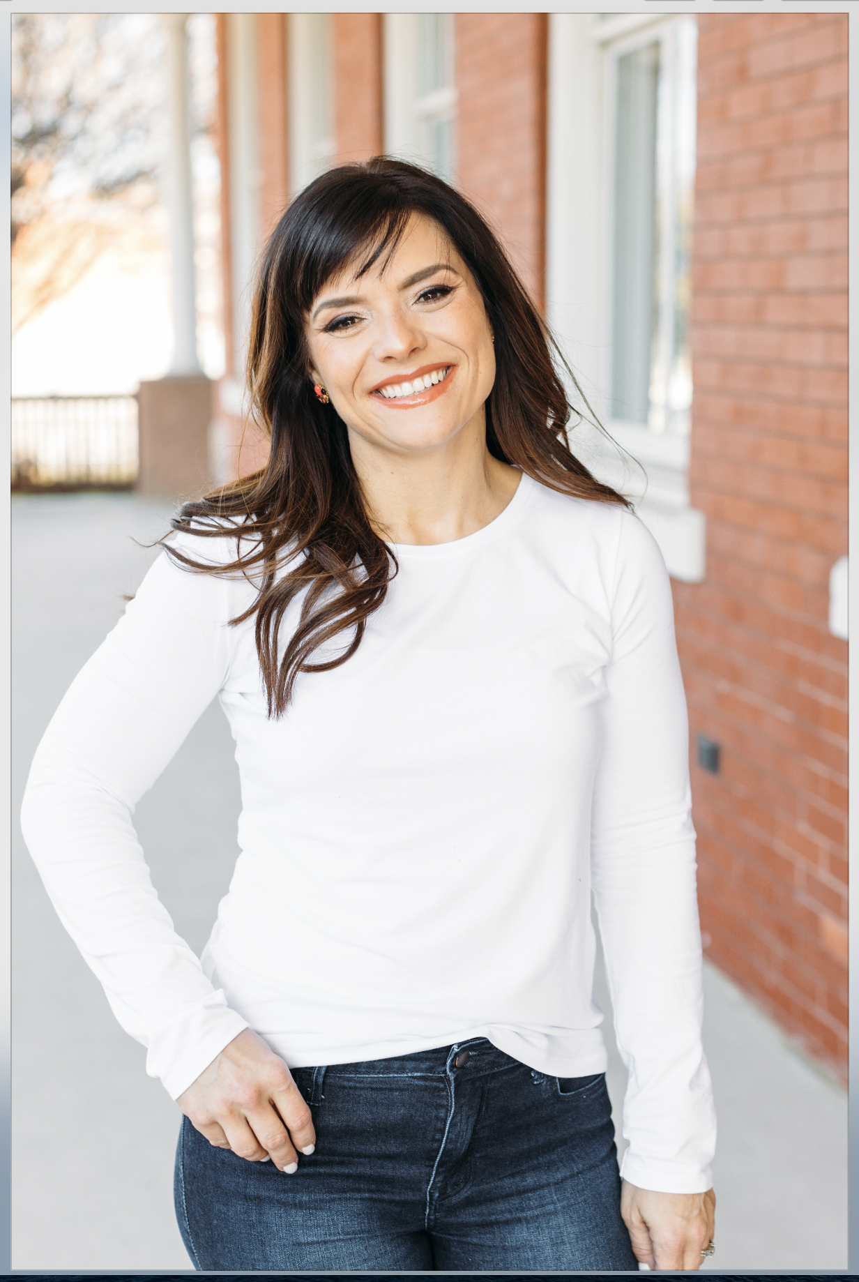 A smiling woman with dark brown hair and makeup, wearing a white long-sleeve shirt and dark jeans, standing outdoors beside a brick building with windows, during daytime.
