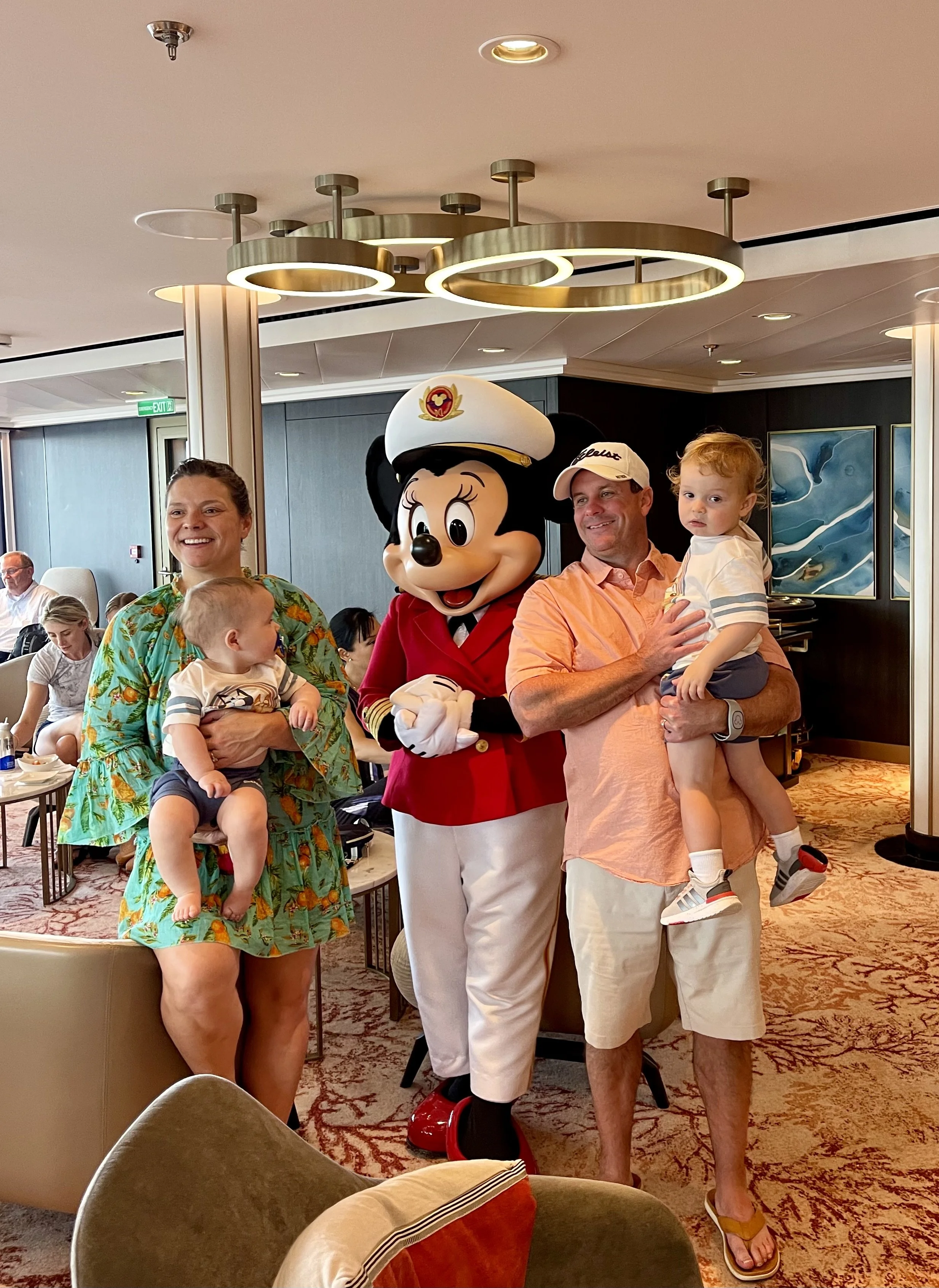A group of people, including a woman, a man, a child, and a person in a Mickey Mouse costume, gathered in a restaurant or hotel lobby. Mickey Mouse is dressed as a captain. The people are smiling and posing for a photo.