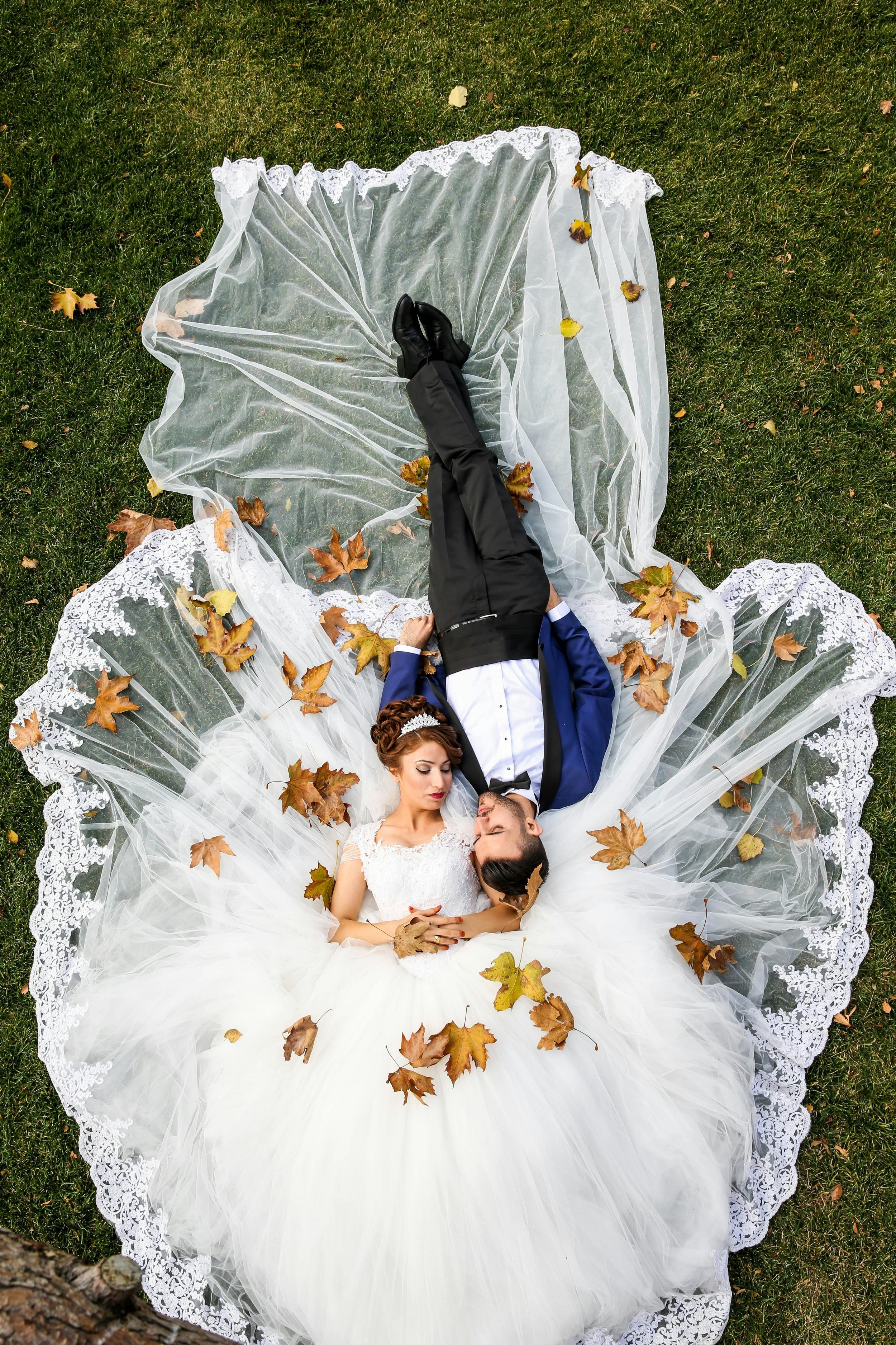 A bride and groom lying on the grass with leaves around them, a lace-trimmed veil spread out beneath them, seen from above.