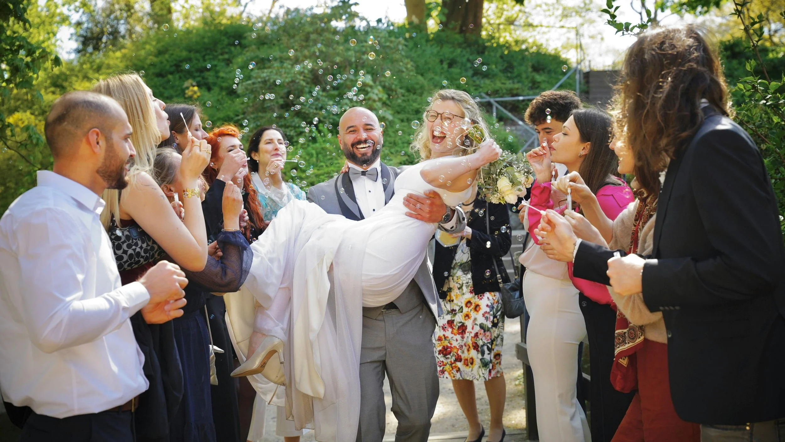A group of people, including a bride and groom, celebrating outdoors with bubbles and smiles.