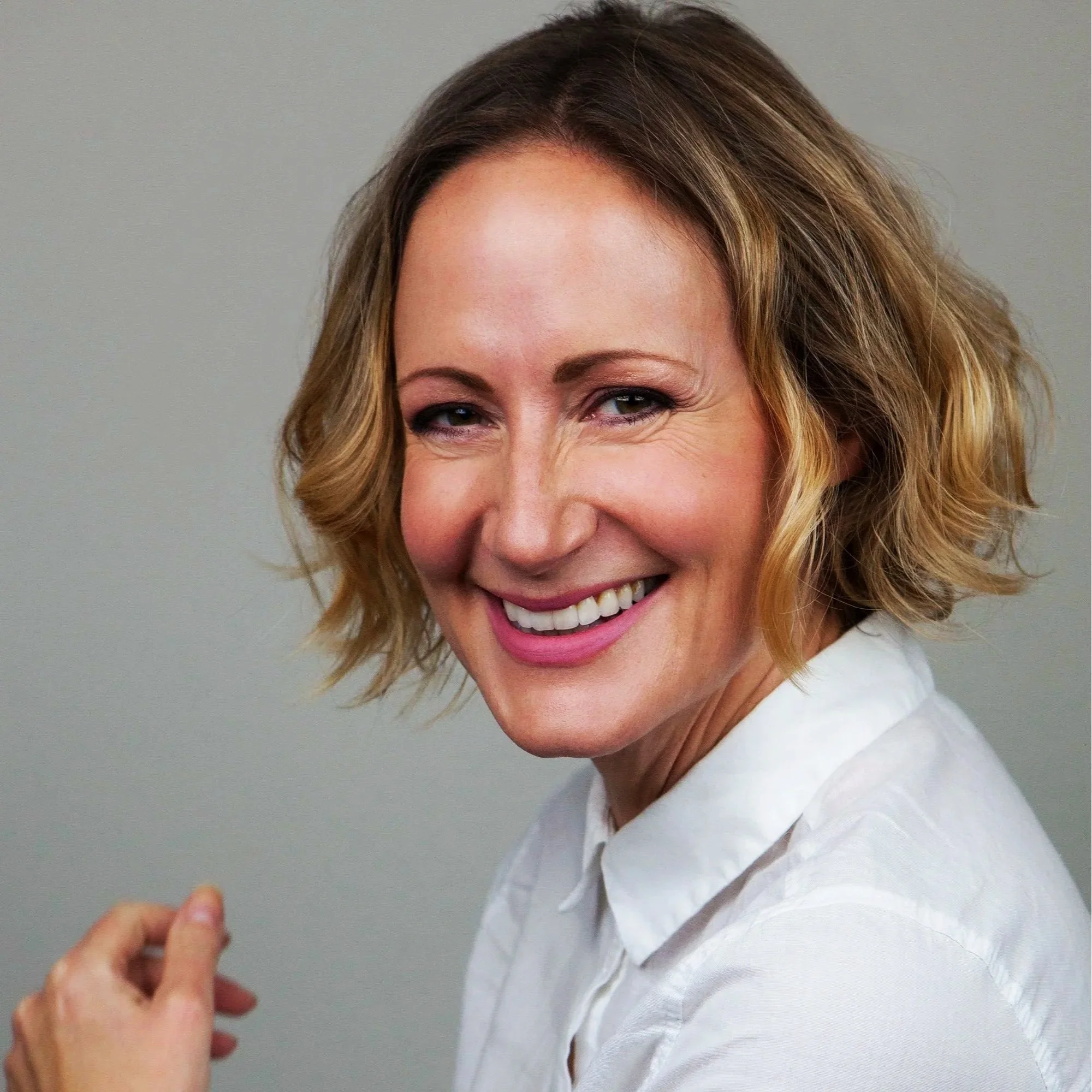 Close-up Kate (blonde, wavy hair smiling, wearing a white blouse against a plain gray background).