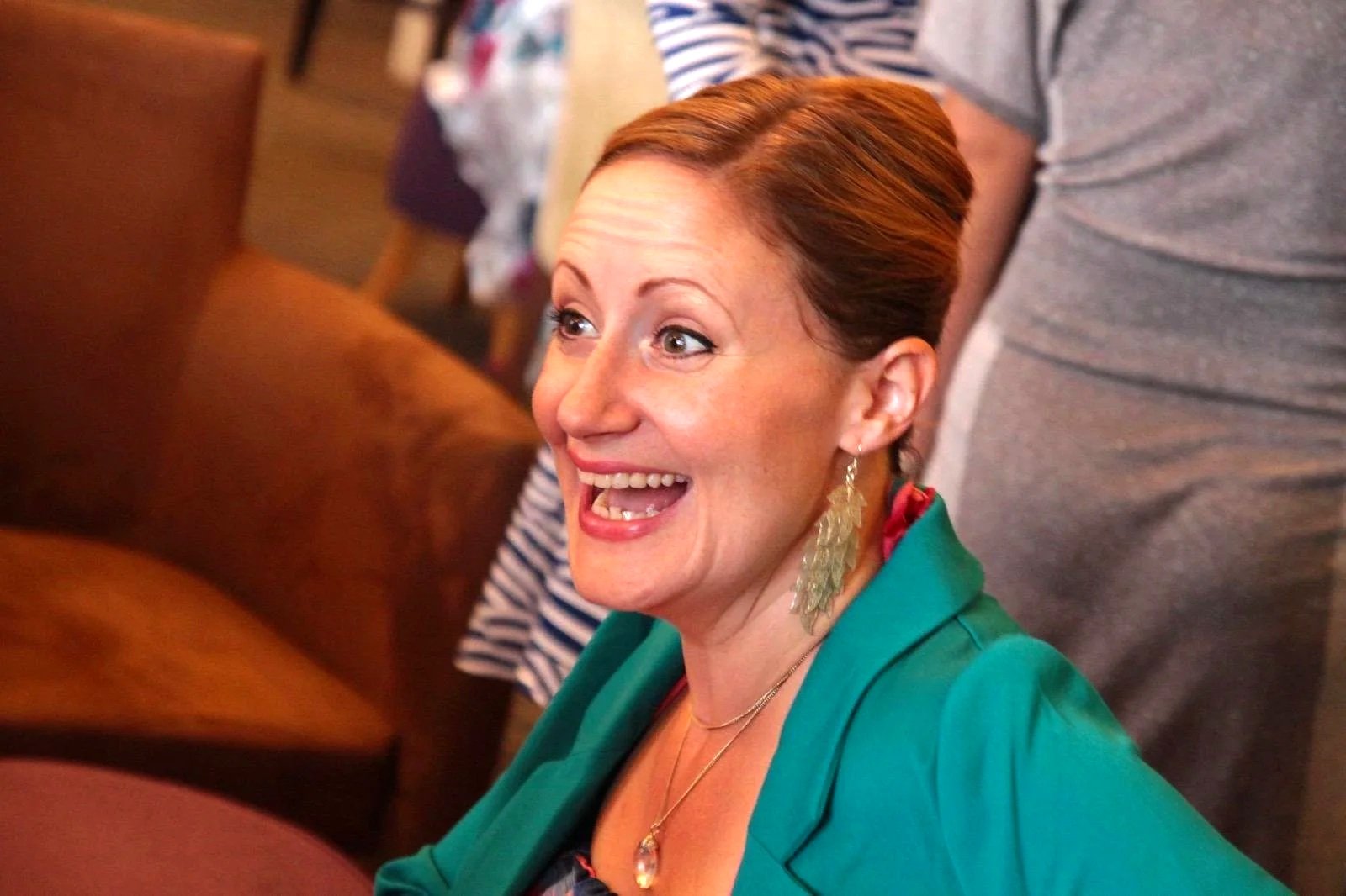A woman with short reddish hair smiling and looking to her left at a social gathering, wearing a teal blazer, gold earrings, and a necklace, with other people in the background.