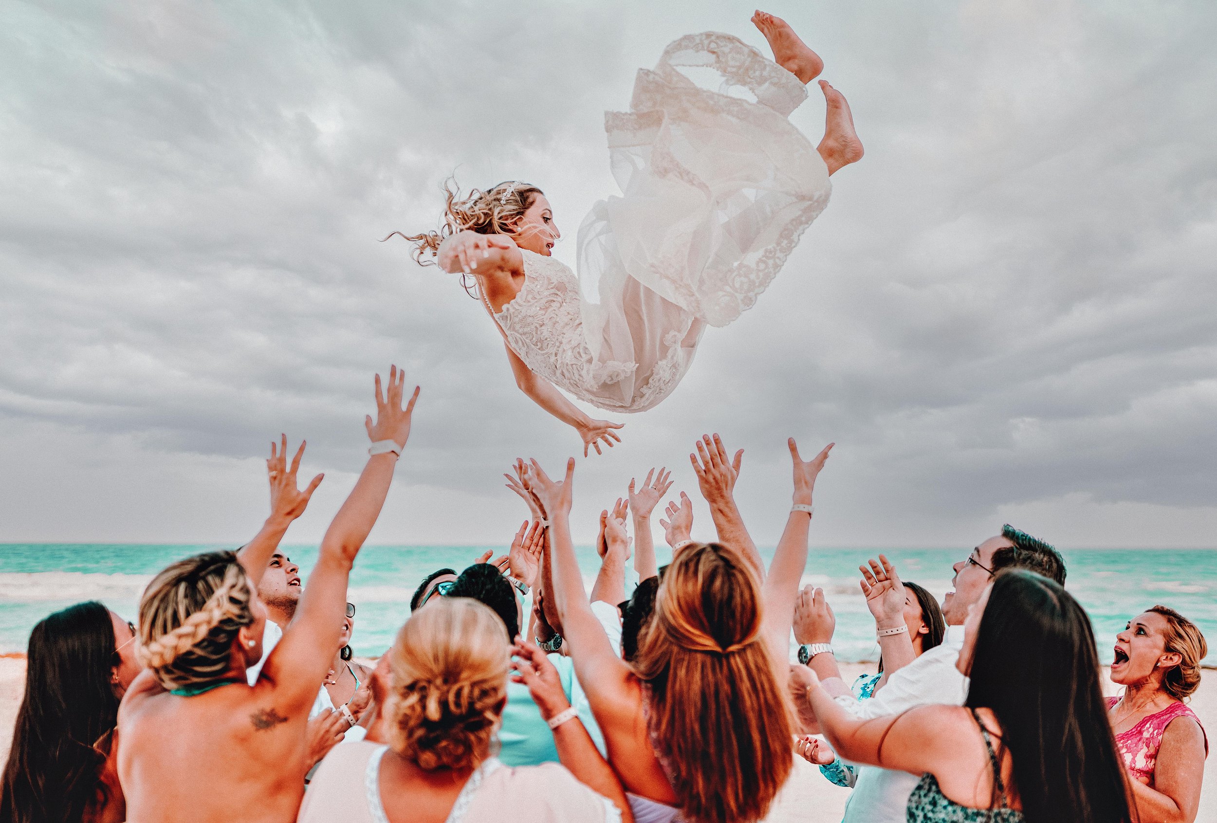 A group of people on a beach lifting a woman in a wedding dress into the air, with the ocean and cloudy sky in the background.