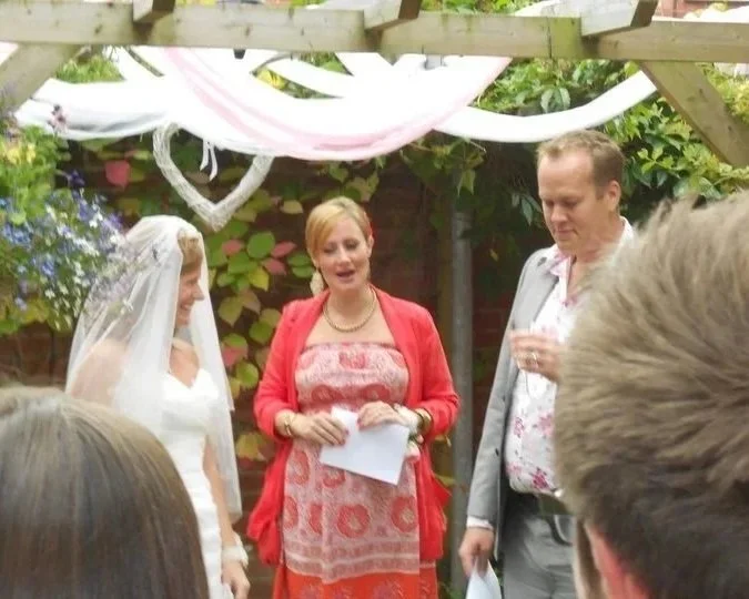 A bride and groom are standing outdoors under a wooden pergola with Kate as a celebrant for their wedding. The grrom is holding a drink. People are gathered around in a garden setting with greenery and flowers.