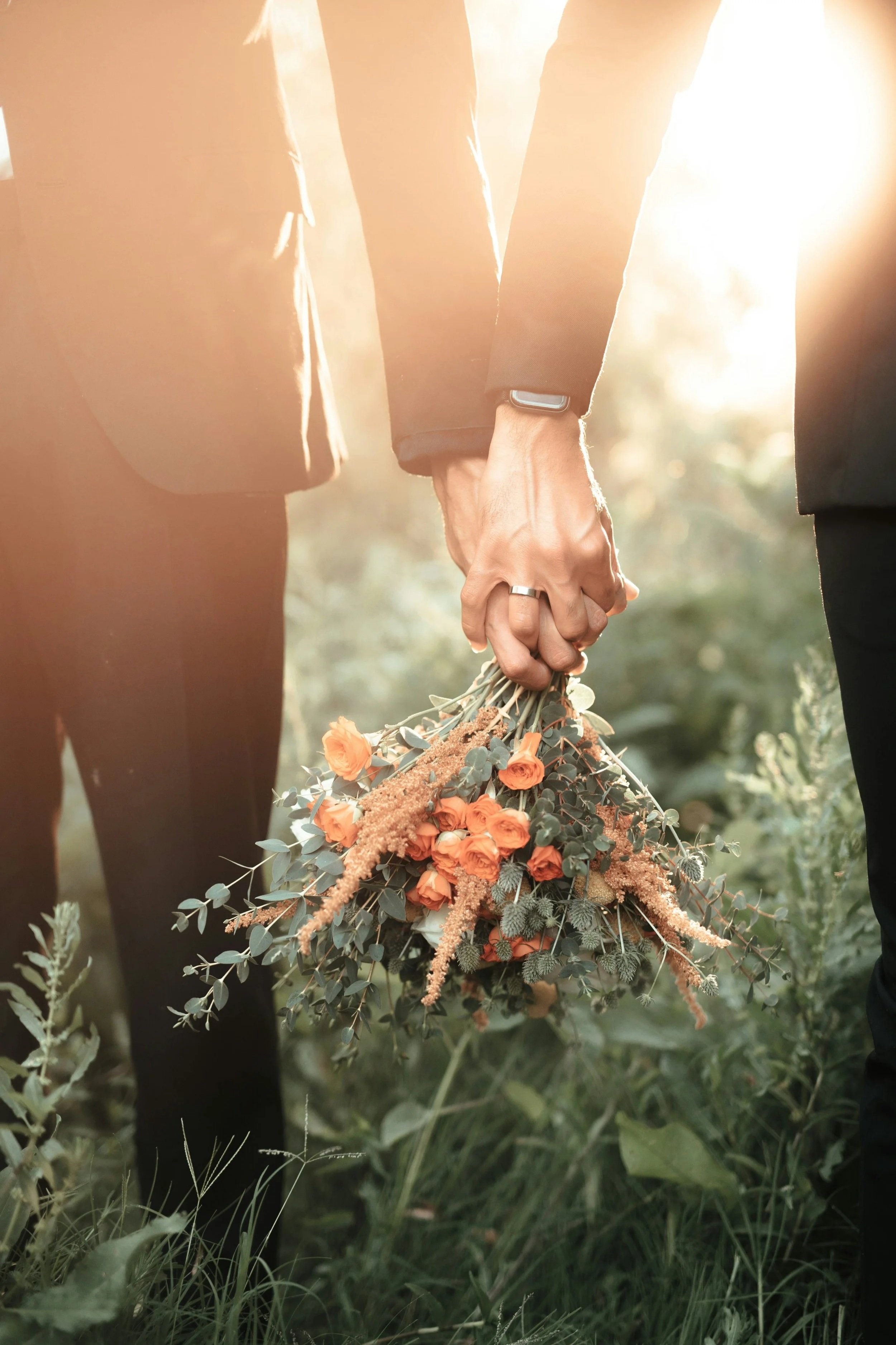 Two people holding hands, one person wearing a smartwatch and ring, with a bouquet of orange flowers and greenery, sunlight shining from behind. The image implies a same sex couple