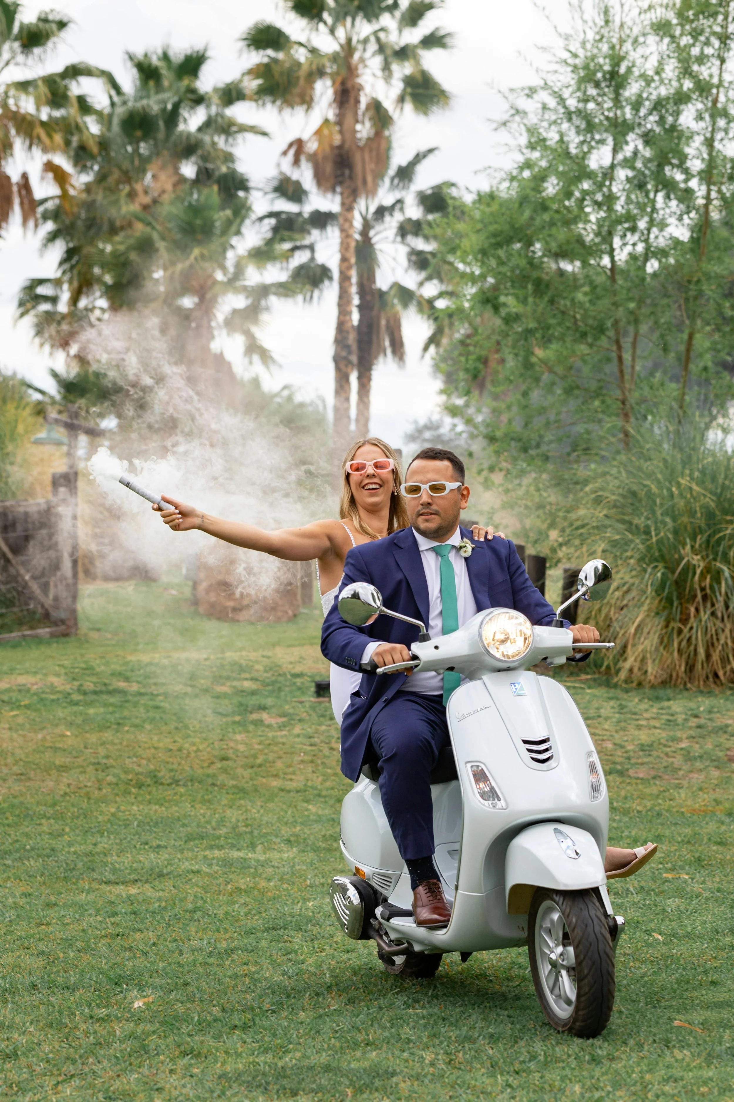 A man in a blue suit and a woman in a white dress riding a white motor scooter through a park with palm trees and greenery. The woman is holding a smoke grenade, creating a cloud of smoke behind her.