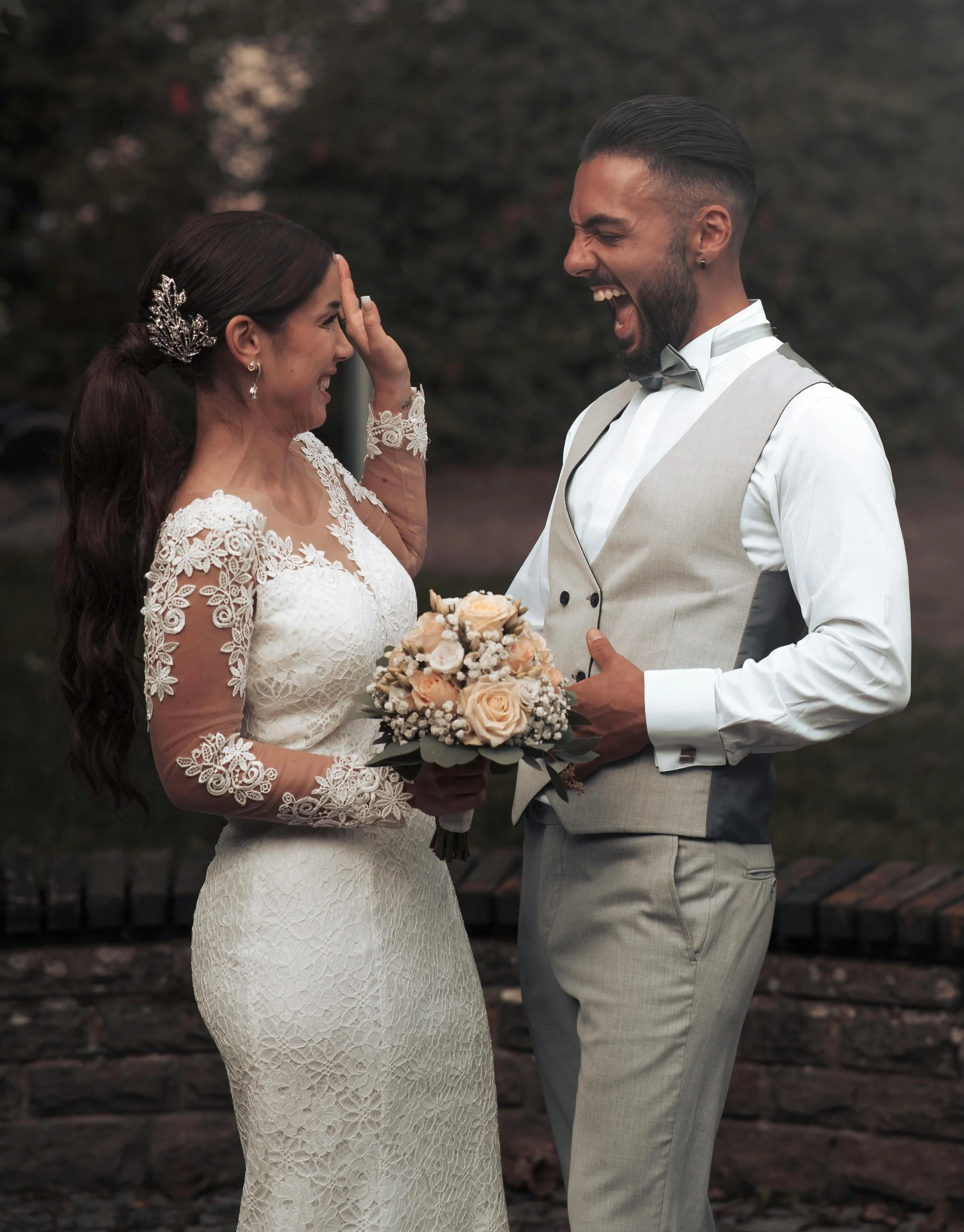 A bride and groom laughing and smiling during their wedding ceremony outdoors, with the bride in a lace wedding dress holding a bouquet of flowers and the groom in a light suit with a bowtie.