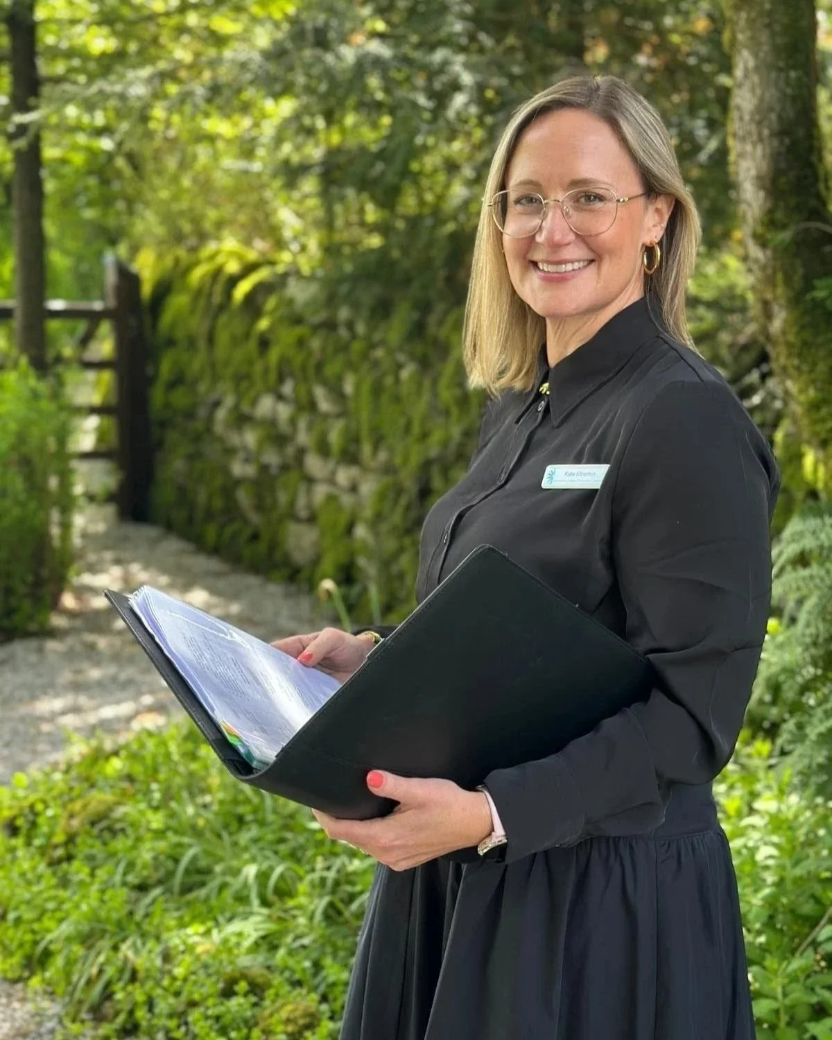 A woman (Kate) with glasses and a black dress standing outdoors with trees and greenery, holding a folder with papers and smiling at the camera.
