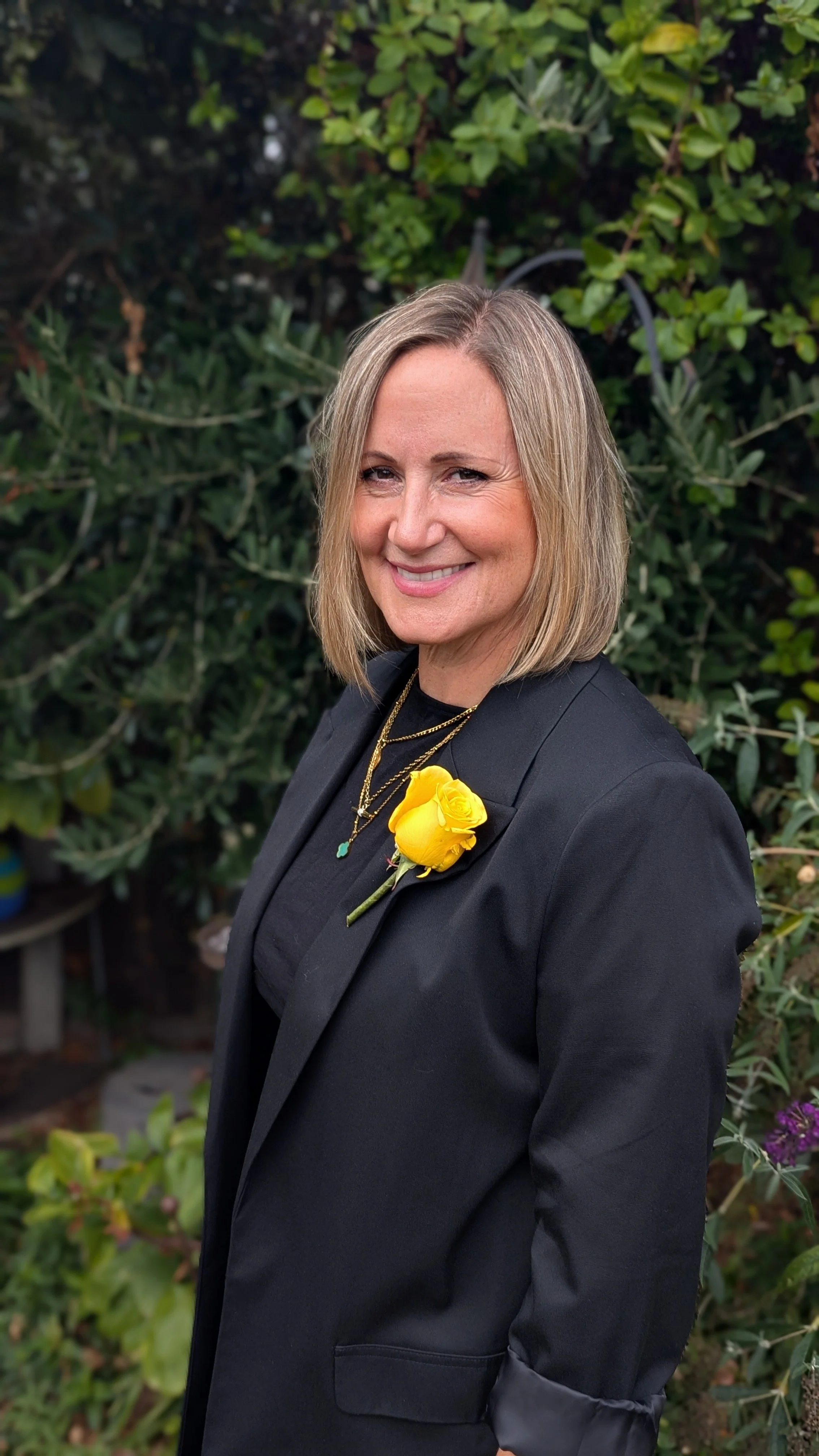 A woman with blonde hair wearing a black blazer, gold necklaces, and a yellow flower on her blazer, standing outdoors in front of green foliage, smiling at the camera.