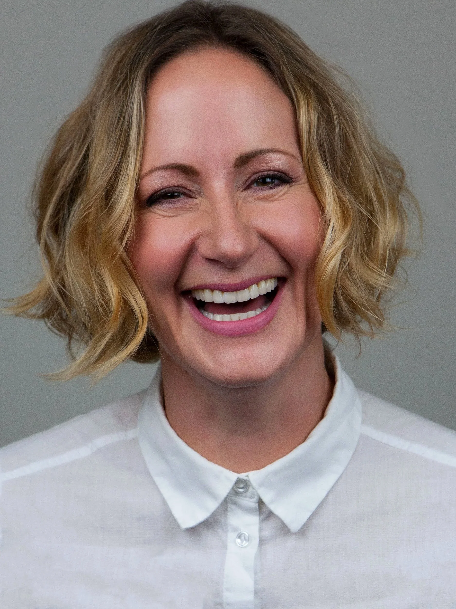 Close-up of Kate (blonde, wavy hair, smiling and showing her teeth), wearing a white collared shirt against a plain grey background.