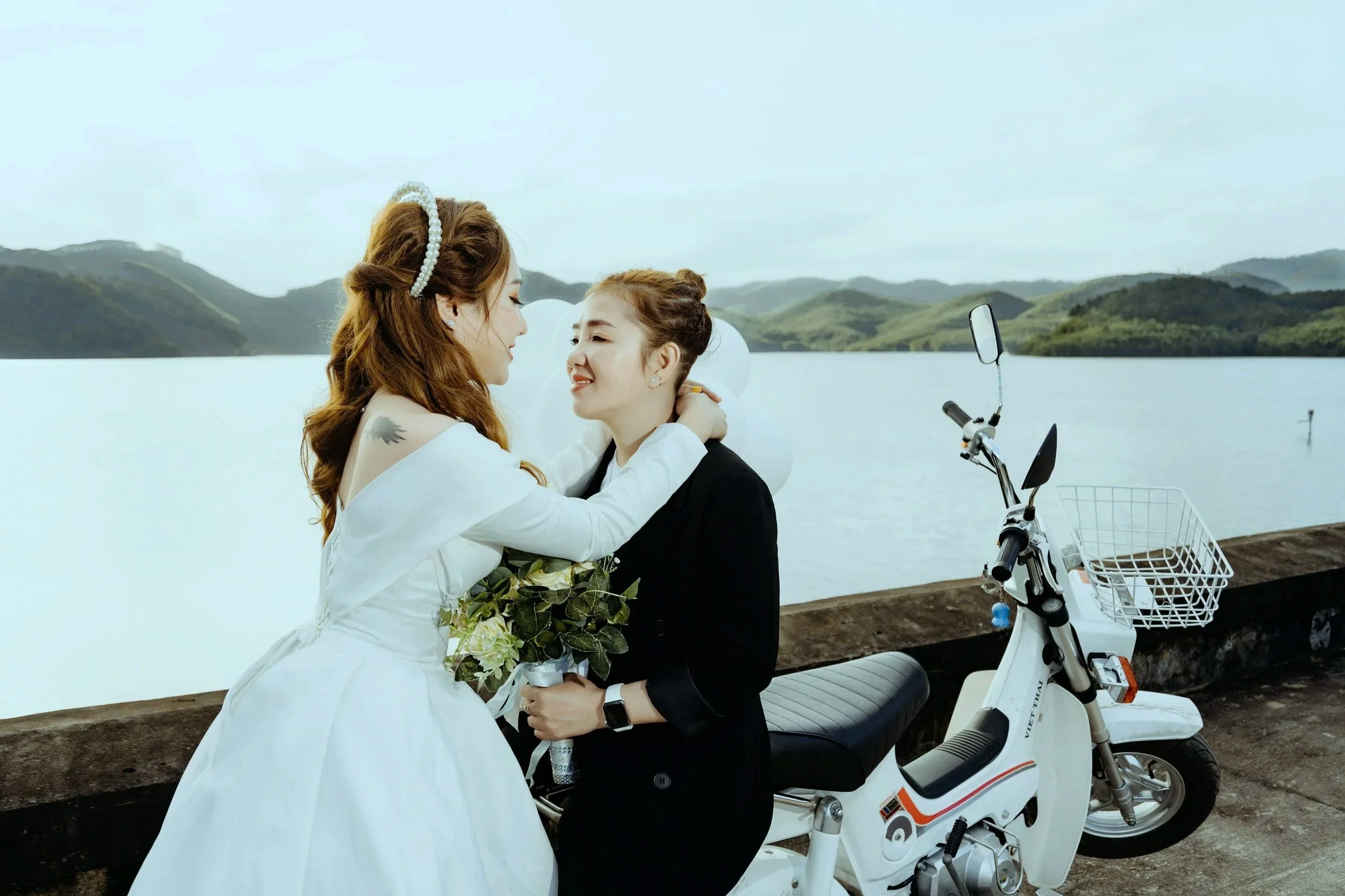 Two women, one dressed in a white wedding gown and the other in a black outfit, sharing an intimate moment outdoors near a body of water with mountains in the background. A white electric scooter is parked nearby.