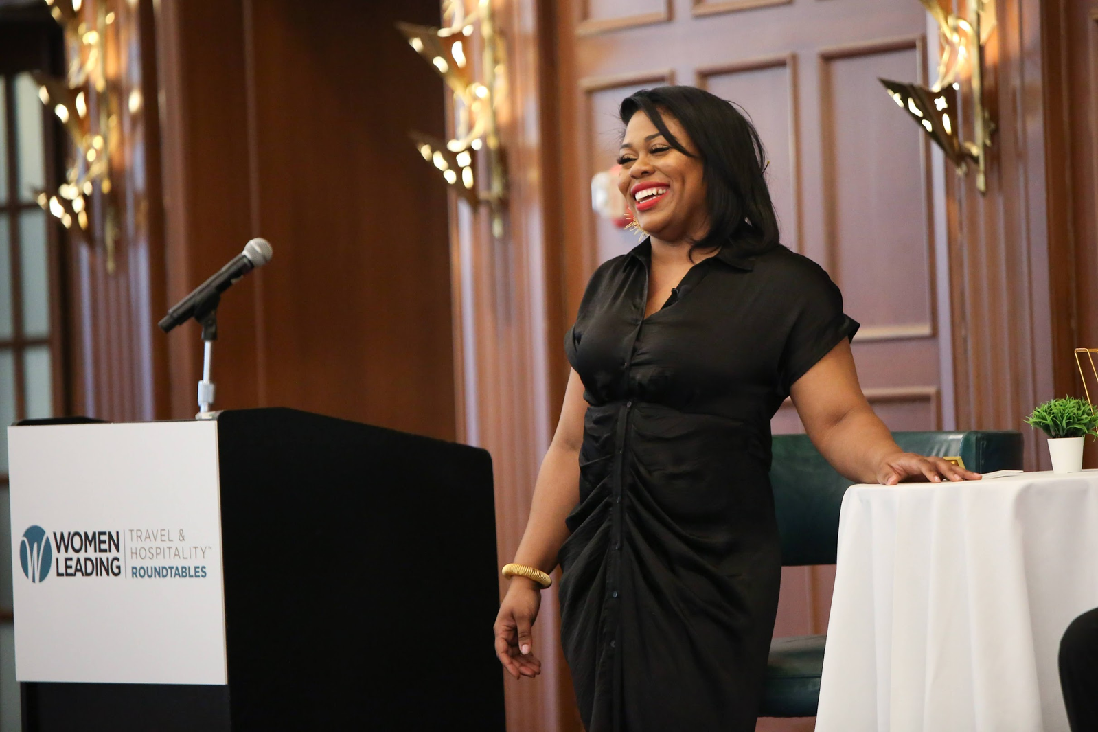 A woman in a black dress standing in front of a podium with a microphone, smiling during a conference or event, with a sign on the podium that reads 'Women Leading Travel & Hospitality Roundtables'.