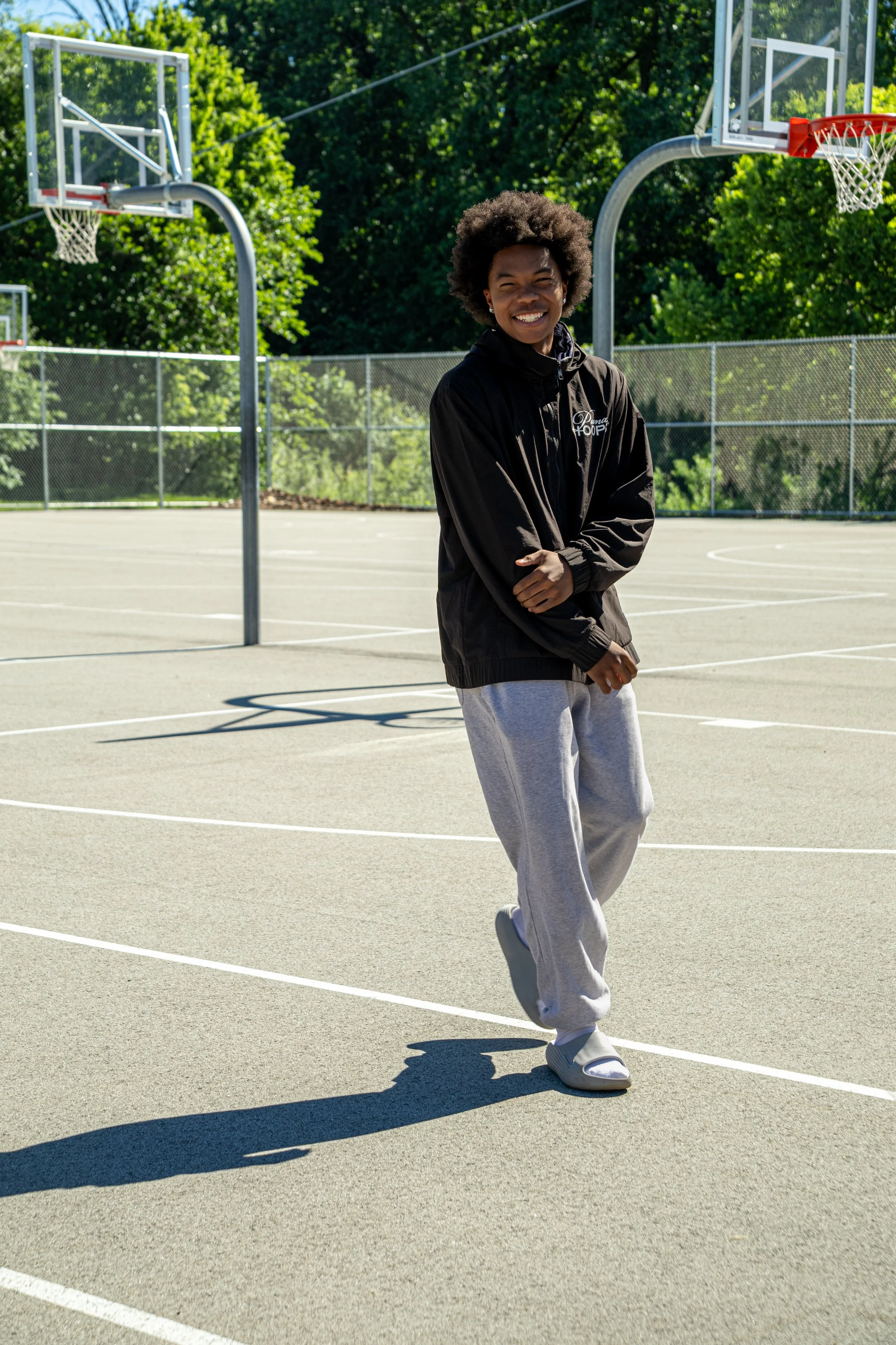 A young woman smiling and walking on an outdoor basketball court, wearing a black jacket, gray sweatpants, and white sneakers. Several basketball hoops and a chain-link fence are visible, surrounded by green trees.