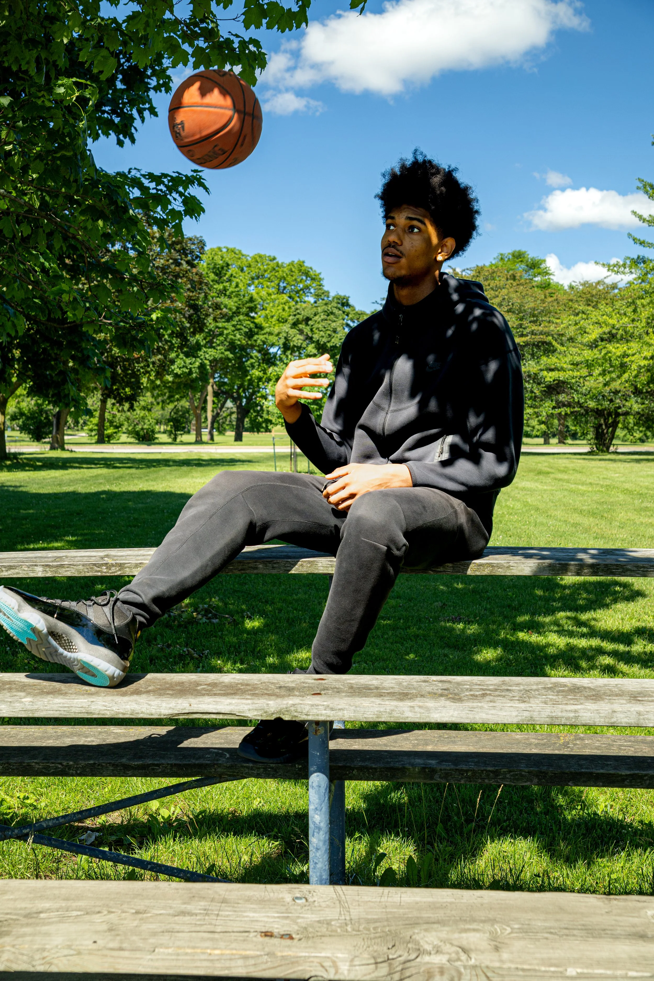 A young man with curly black hair, sitting on a wooden bench outdoors, watching a basketball in the air, with a background of green trees and a partly cloudy blue sky.