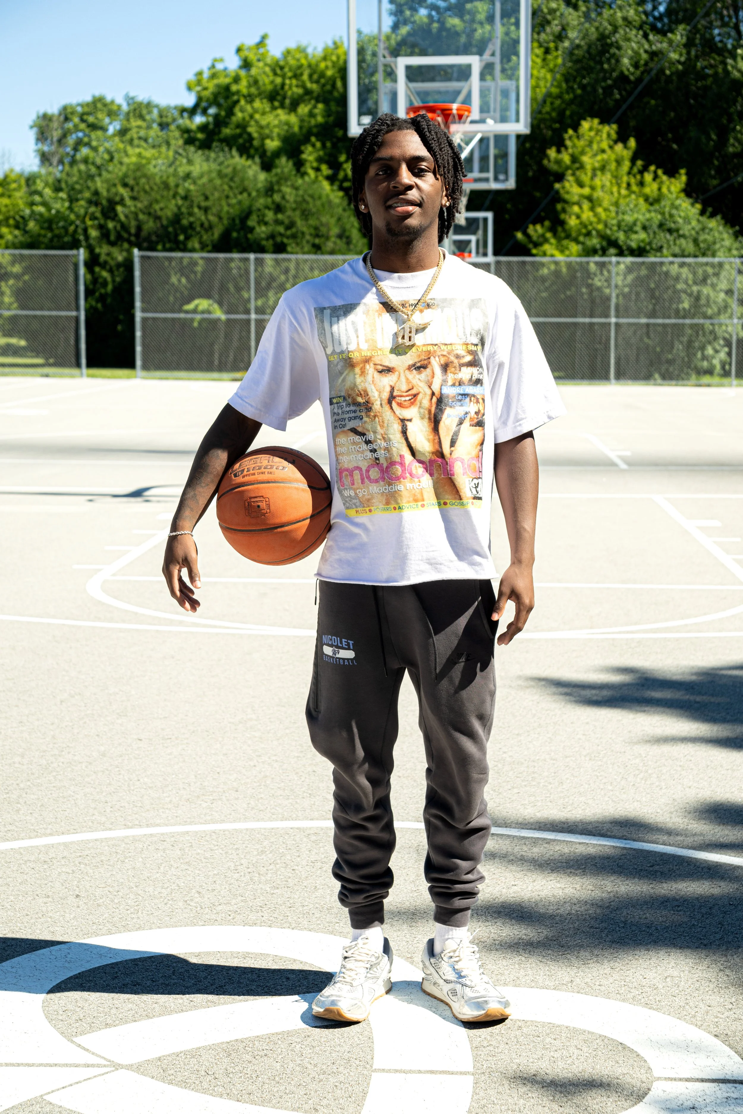 Young man standing on an outdoor basketball court holding a basketball, wearing a white Madonna graphic T-shirt, gray sweatpants, and white sneakers, with a basketball hoop and green trees in the background on a sunny day.