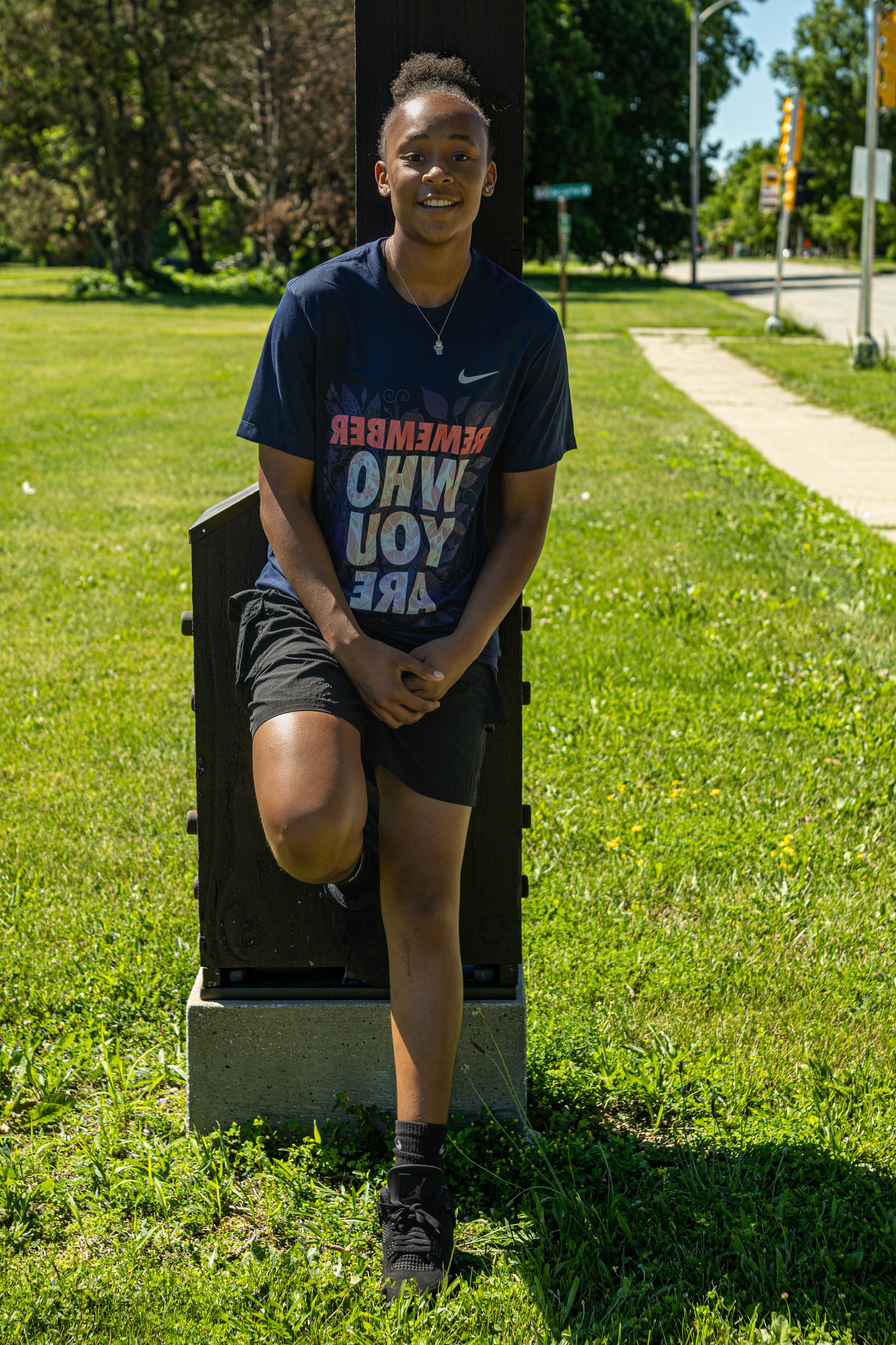 A young girl with a natural hairstyle leaning against a wooden post outdoors on a sunny day, wearing a dark t-shirt and shorts, with greenery and a sidewalk in the background.