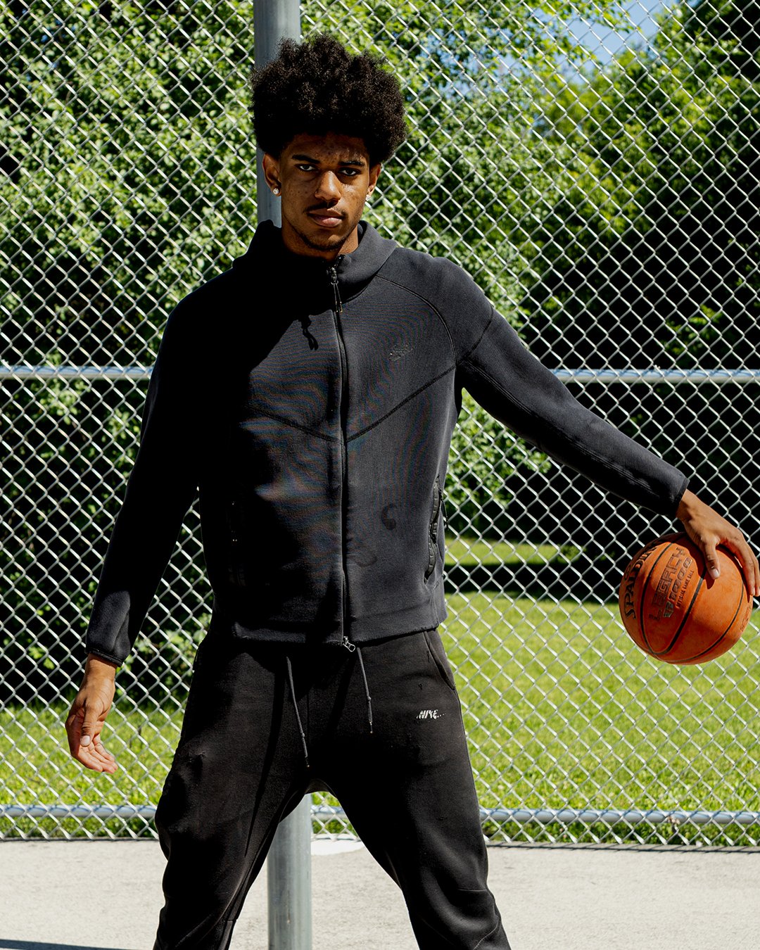 Young man holding basketball on outdoor tennis or basketball court with chain-link fence and green trees in background.