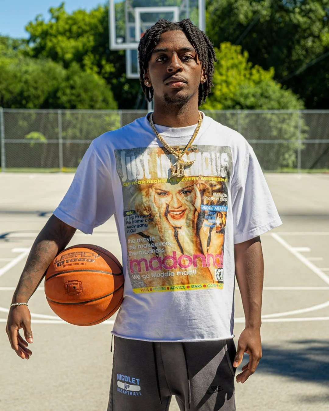 Young man standing on an outdoor basketball court, holding a basketball, wearing a vintage Madonna T-shirt, gold chains, and Nike sweatpants, with trees and a basketball hoop in the background.