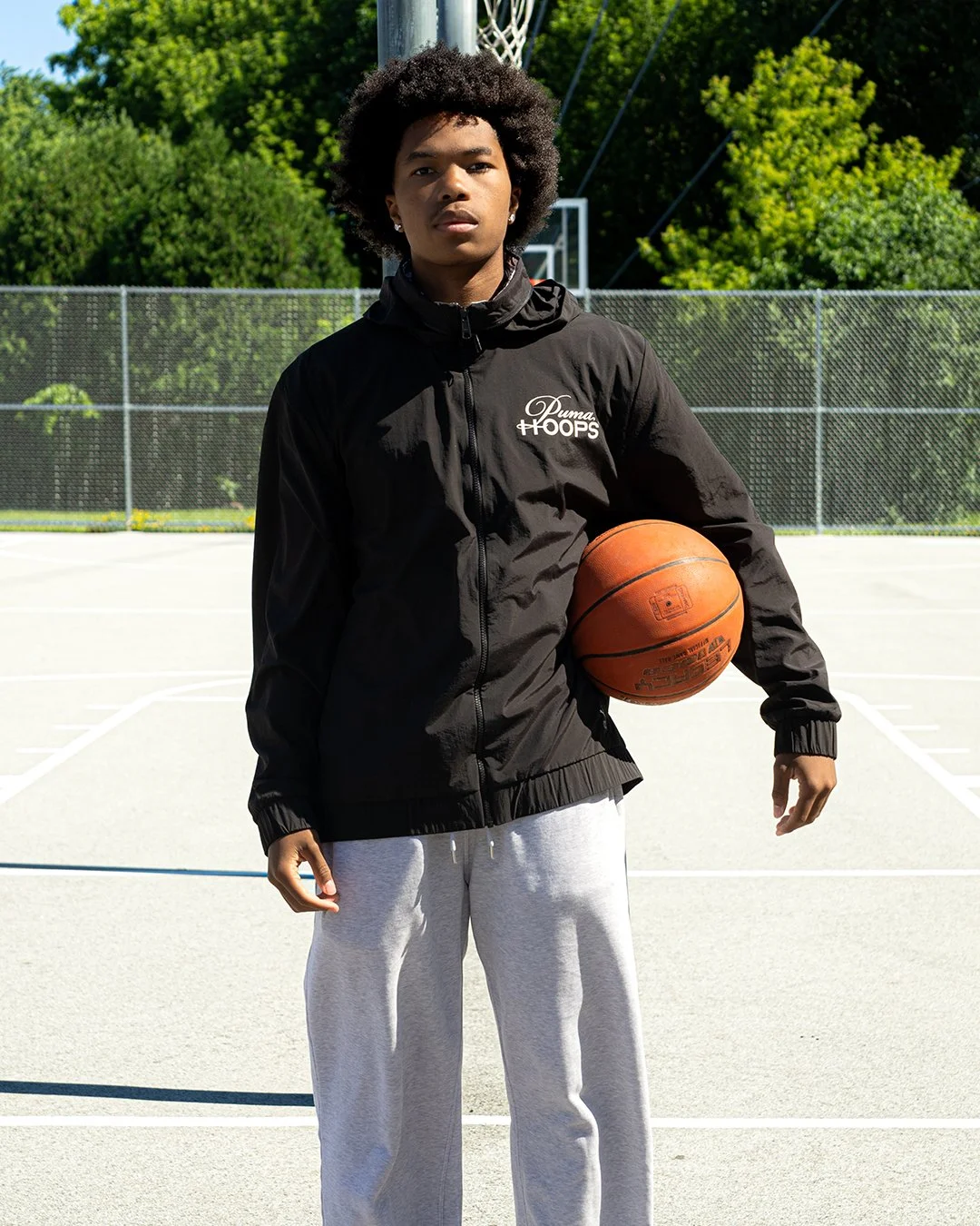 A young man standing on an outdoor basketball court holding a basketball in his right hand, wearing a black Puma Hoops jacket and light gray sweatpants, with trees and a chain-link fence in the background.