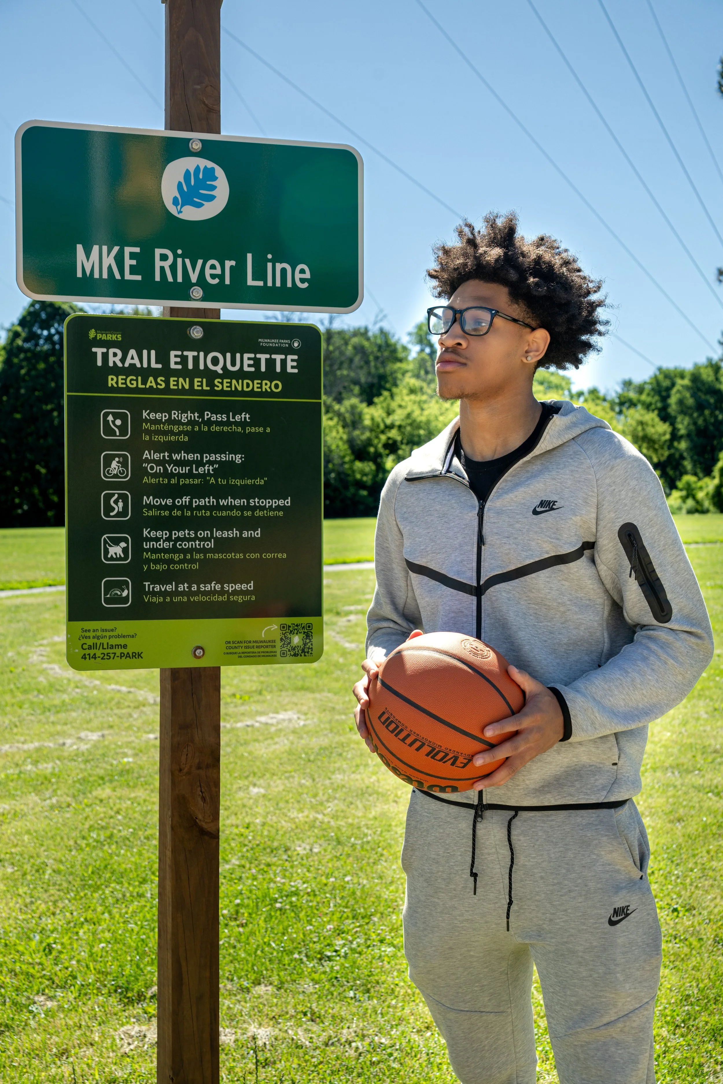 A young man holding a basketball stands next to a trail etiquette sign at a park. The sign provides rules for trail use in both English and Spanish, and is mounted on a wooden post. The park has green grass and trees in the background, along with a clear blue sky.