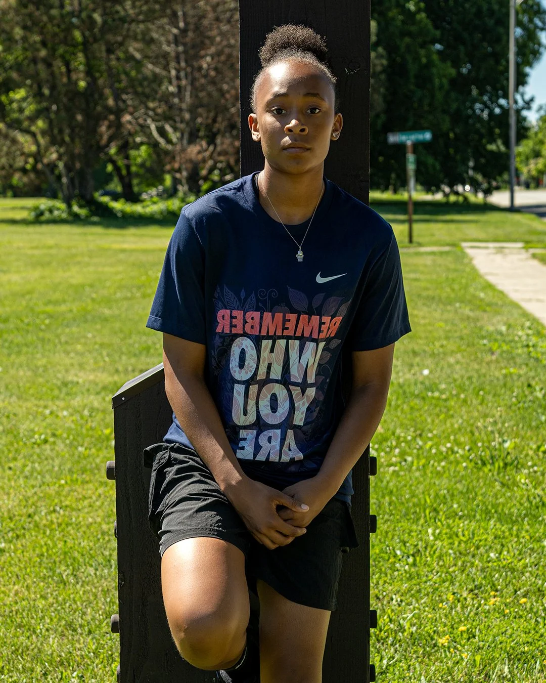 Young woman wearing a black T-shirt with colorful text, black shorts, standing outdoors leaning against a wooden post, with a grassy park and trees in the background.