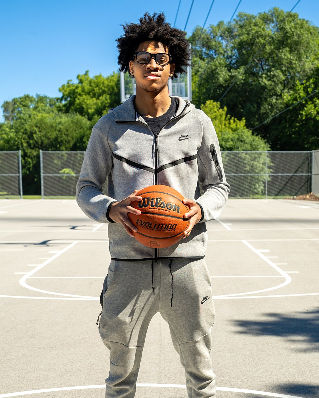 Young man with curly hair and glasses standing on an outdoor basketball court holding a basketball, wearing a gray Nike tracksuit, with green trees and a basketball hoop in the background.