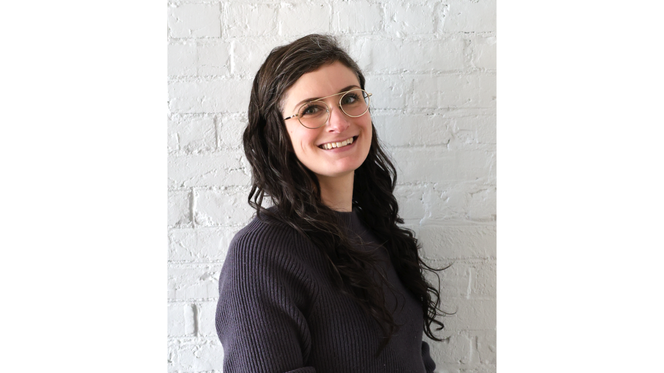 Professional headshot of a smiling woman with long dark hair and glasses, wearing a dark sweater, standing in front of a white brick wall.