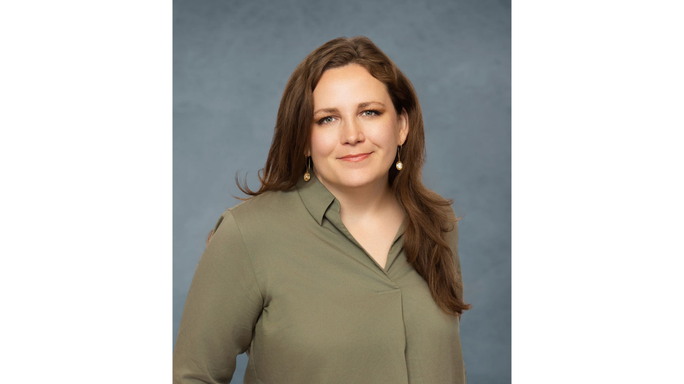 Professional headshot of a woman with long brown hair, wearing a green blouse and earrings, posed against a soft blue-gray background.