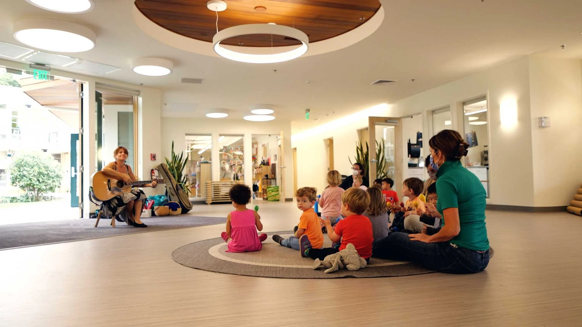 Children and teachers gather for music time in the central piazza, a bright shared learning space with circular lighting, warm wood ceilings, and open connections to the outdoors.