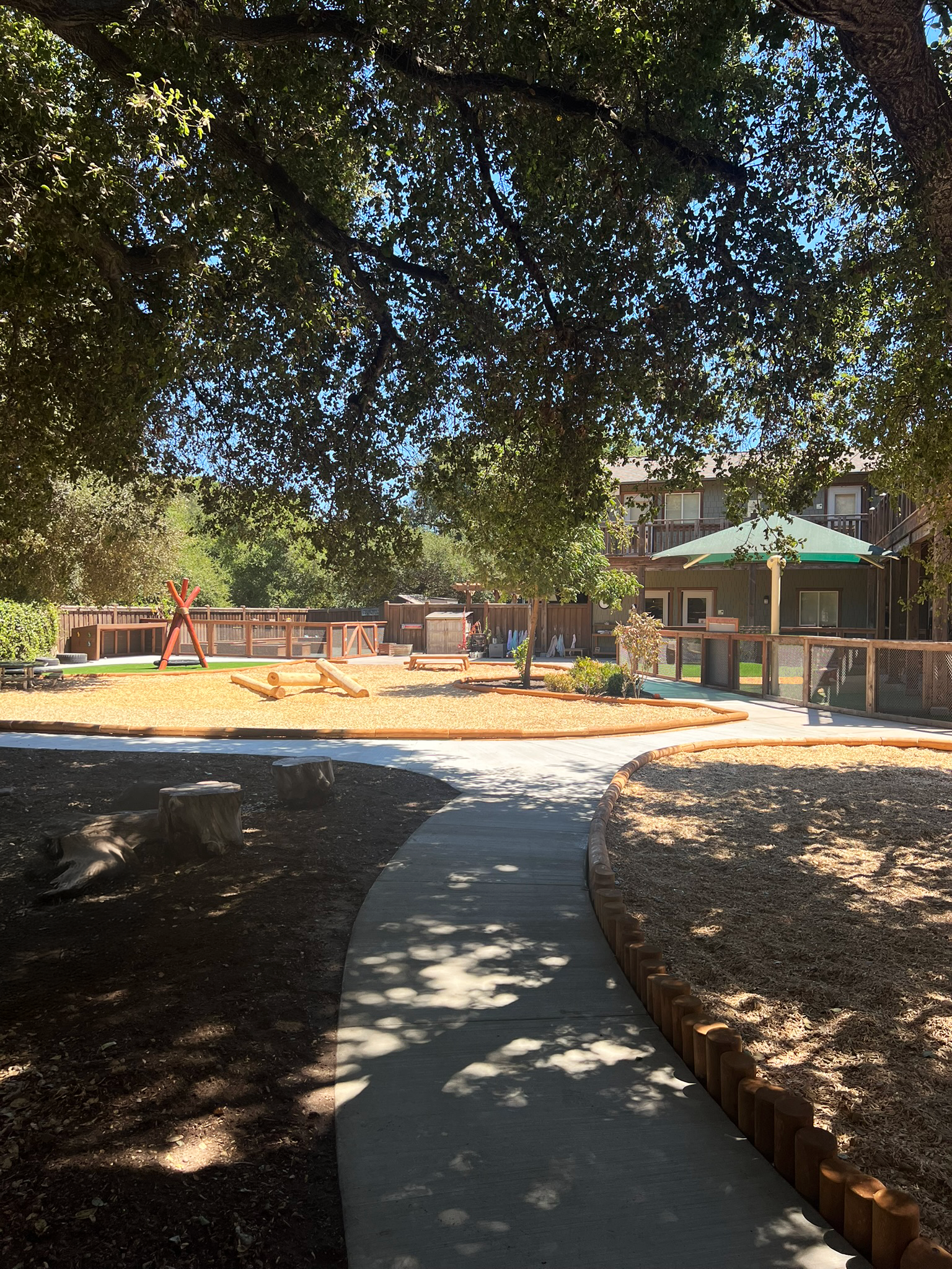 Curving concrete pathway winds beneath mature trees through outdoor play areas with natural wood features and open play spaces.
