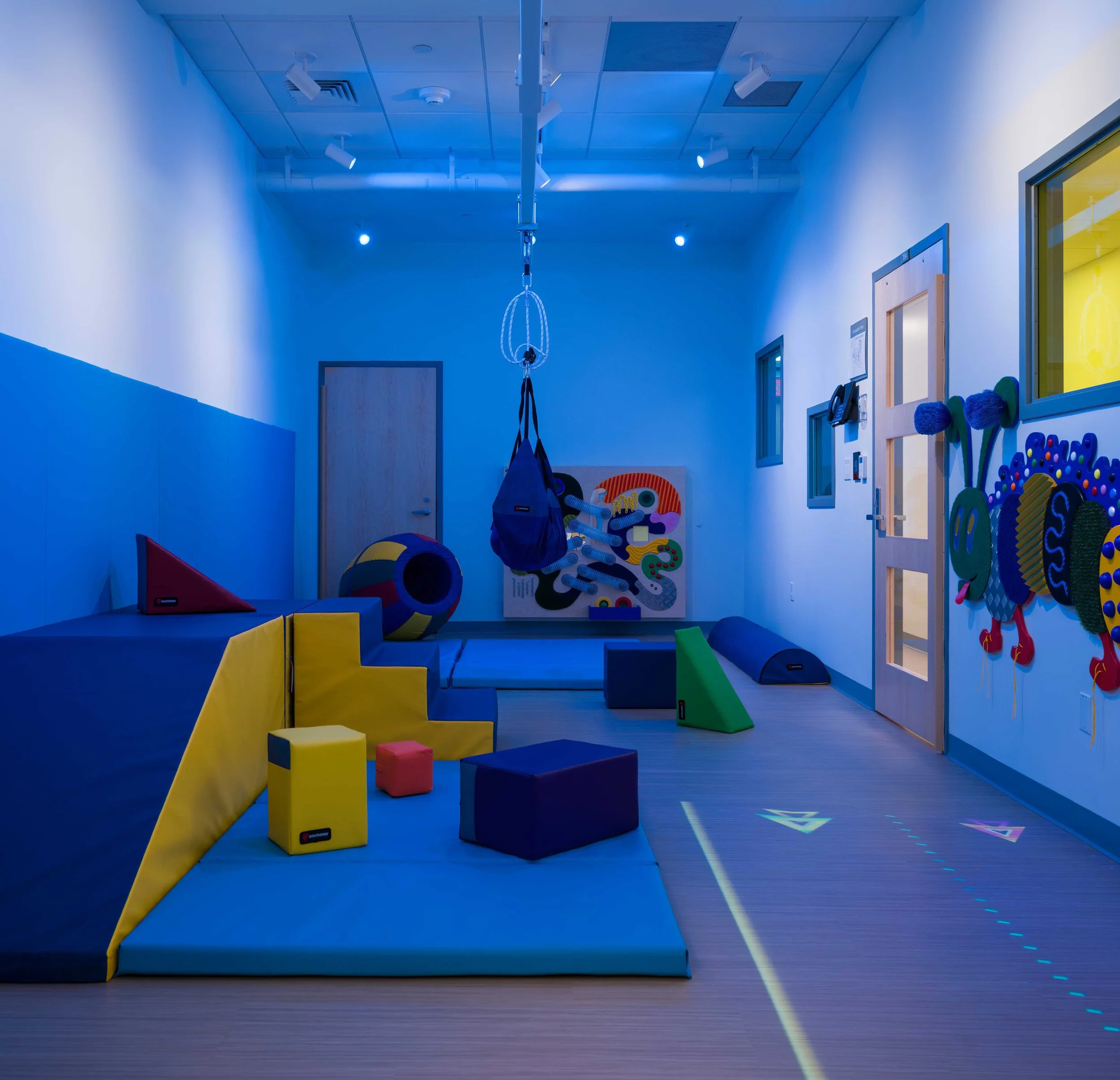 Sensory gross motor room with soft climbing equipment, a suspended sensory swing, interactive wall features, and adjustable lighting to support movement, sensory exploration, and self-regulation.
