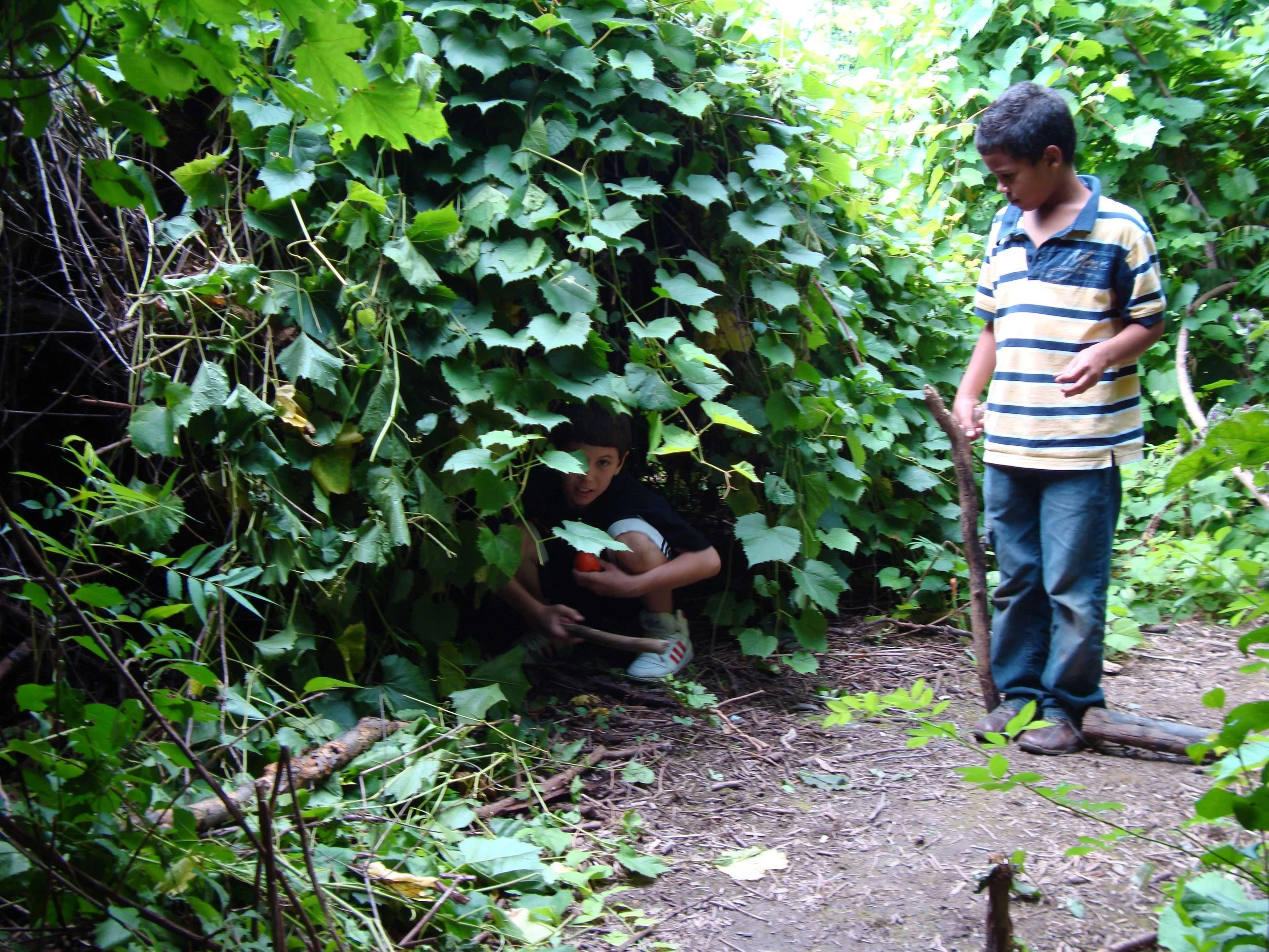 Child crouches inside a leafy, nature-built fort while another child stands nearby on a wooded trail.