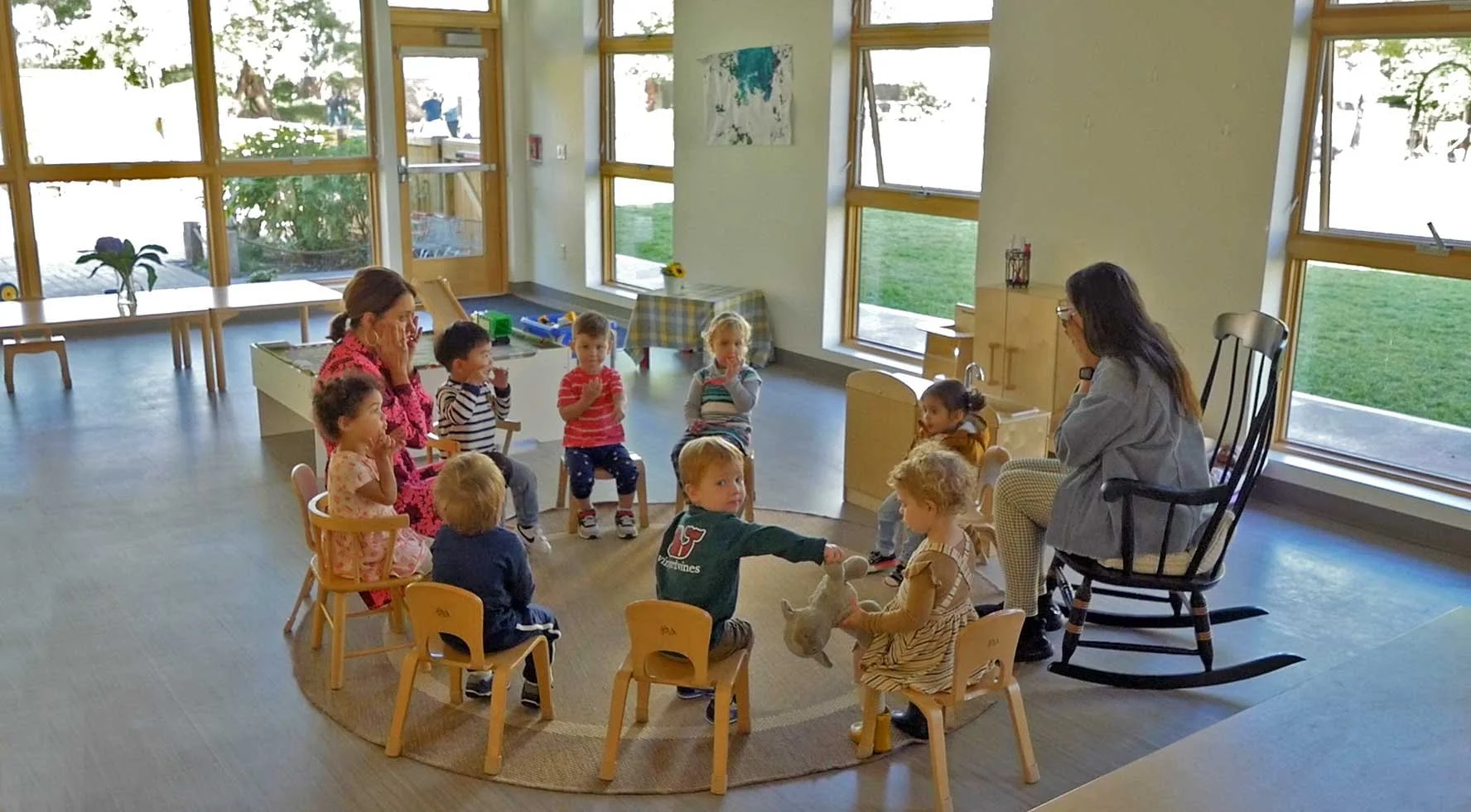 Teachers lead toddlers in circle time inside a sunlit classroom with child-sized furniture, large windows, and direct views of the surrounding natural landscape.