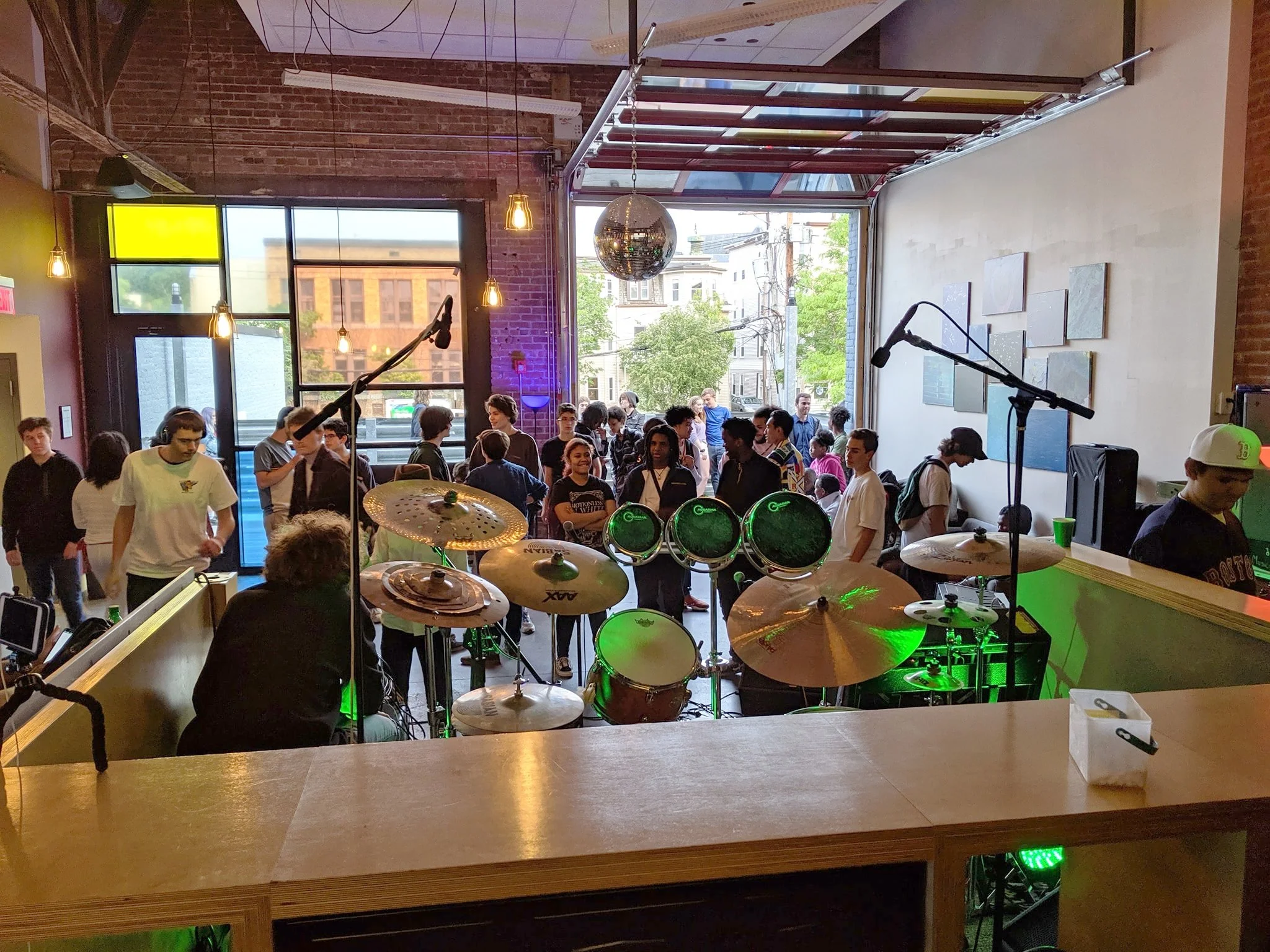 Teens gather around a drum kit and performance setup in a flexible music venue with an open garage-style door.