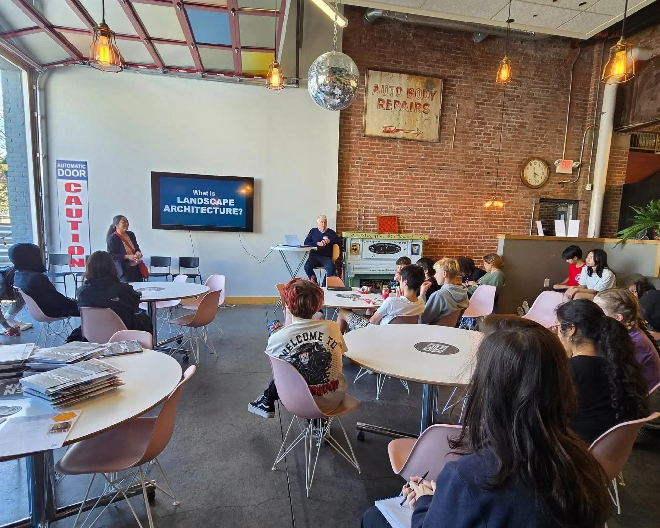 Teens attend a presentation about landscape architecture at the Brookline Teen Center, seated at round tables in a flexible gathering space with exposed brick walls and an open garage door.