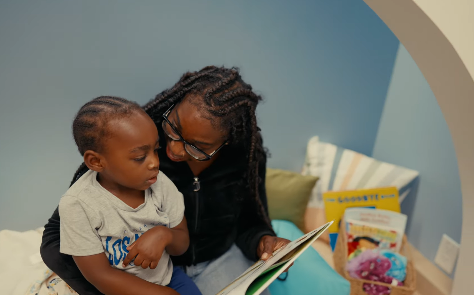 An educator reads with a young child in a semi-enclosed reading nook featuring soft seating, built-in shelving, and a calming blue backdrop.