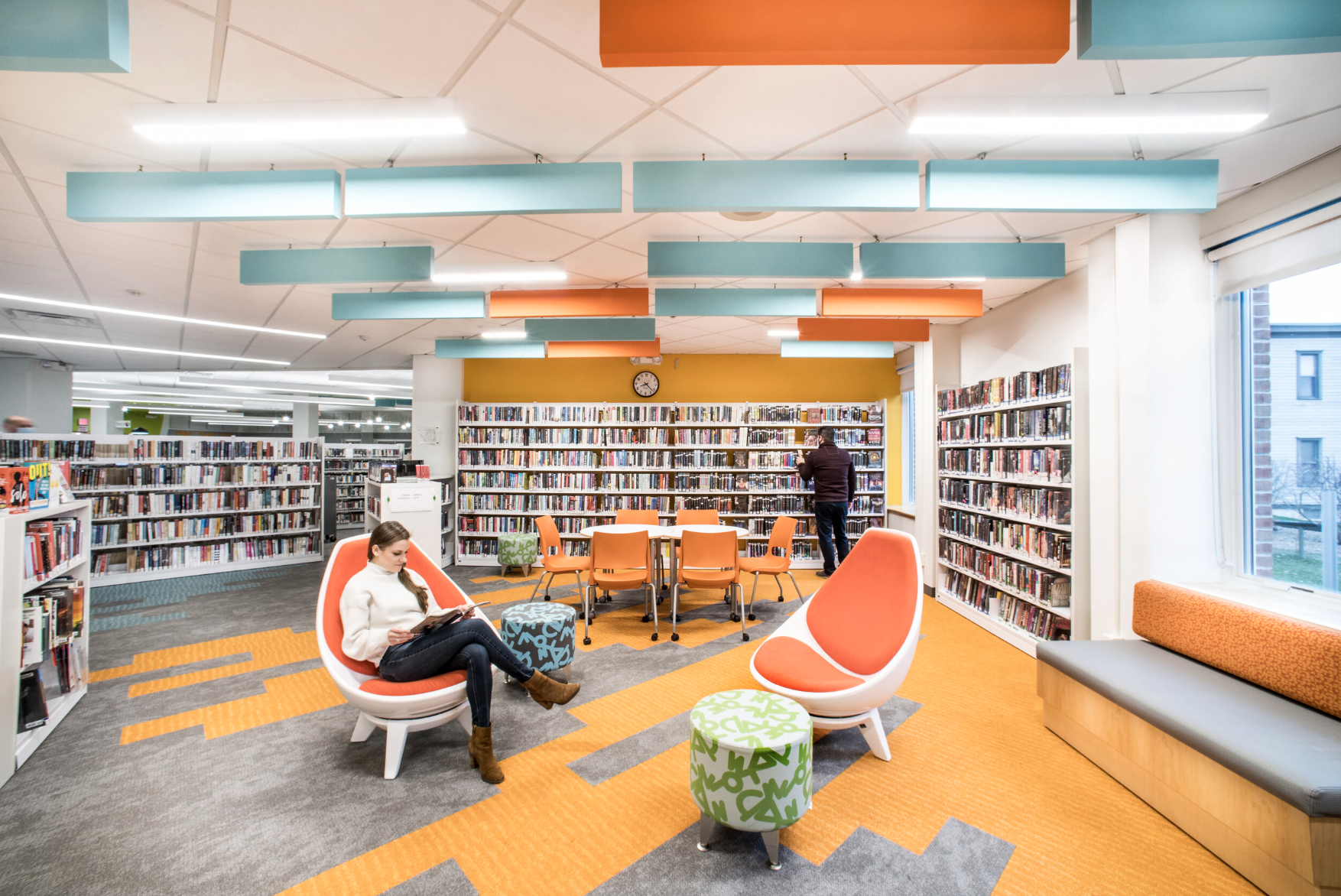 Interior of a modern library with orange and teal decor, bookshelves filled with books, and four people reading or browsing.