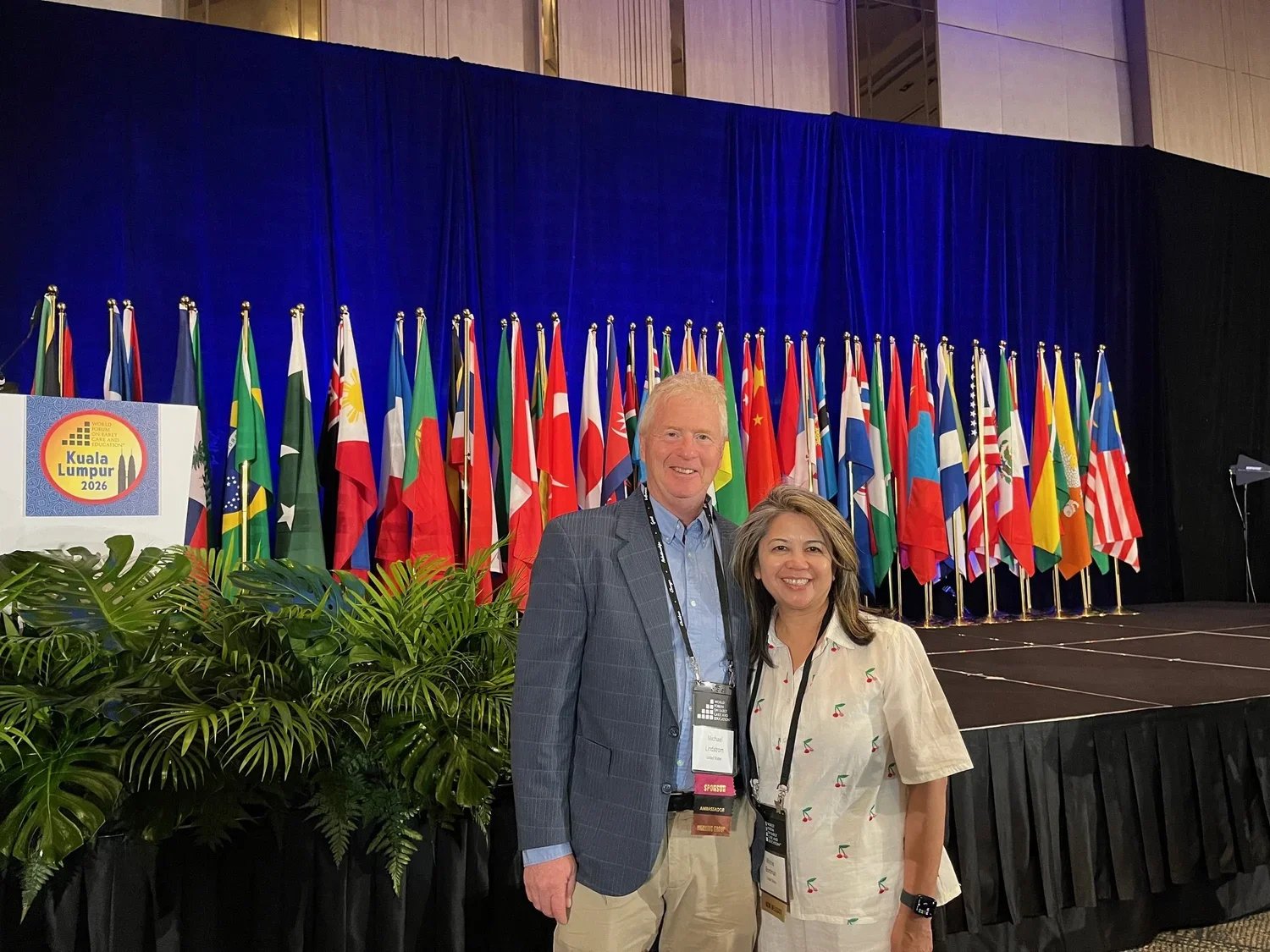 Two conference attendees pose for a photo in front of a stage lined with international flags. A Kuala Lumpur 2026 conference sign and tropical plants frame the event setting.