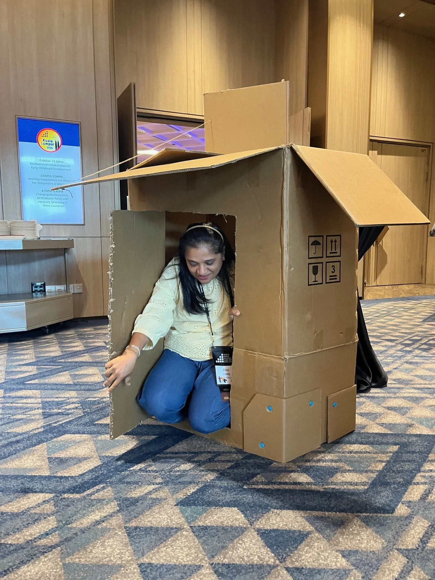 A conference attendee kneels inside a cardboard playhouse during a hands-on workshop activity. The temporary structure features a pitched roof, open doorway, and folded cardboard panels.
